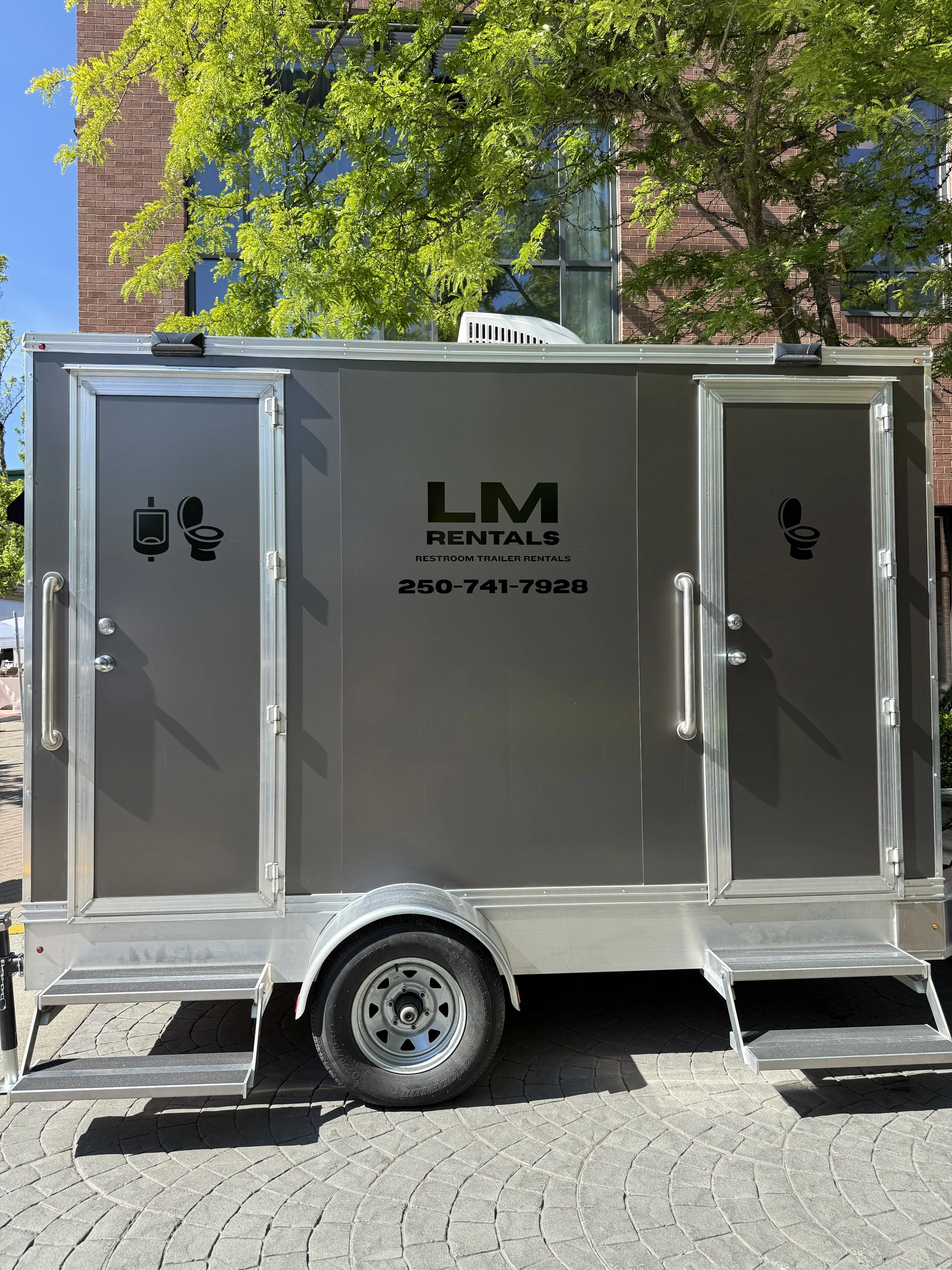 Gray restroom trailer with black company logo and phone number, two steps leading up to doors with toilet icons, parked outdoors.