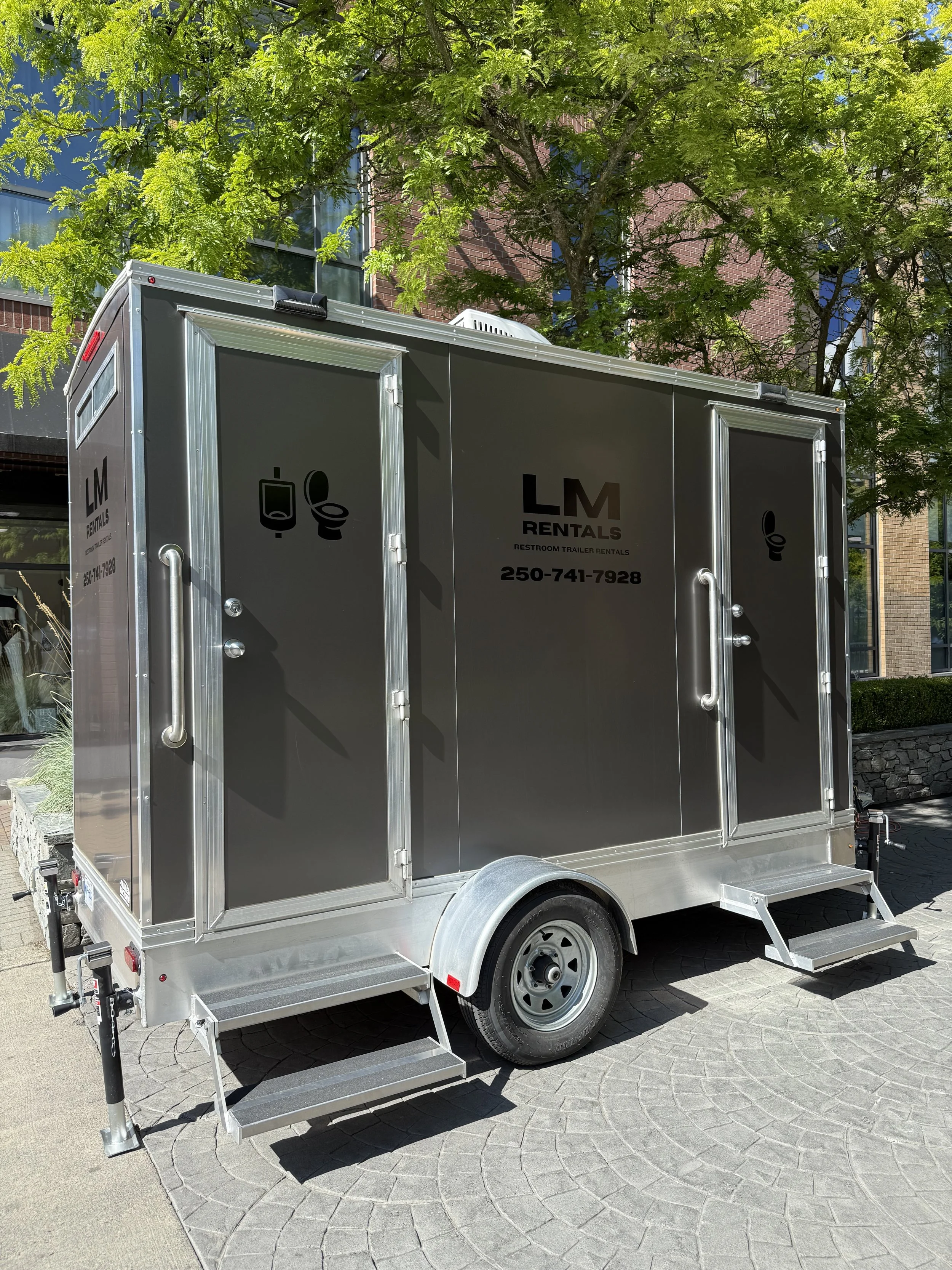 Restroom trailer with two doors, each with a toilet icon, parked on a brick-paved area under a green leafy tree, with buildings in the background.