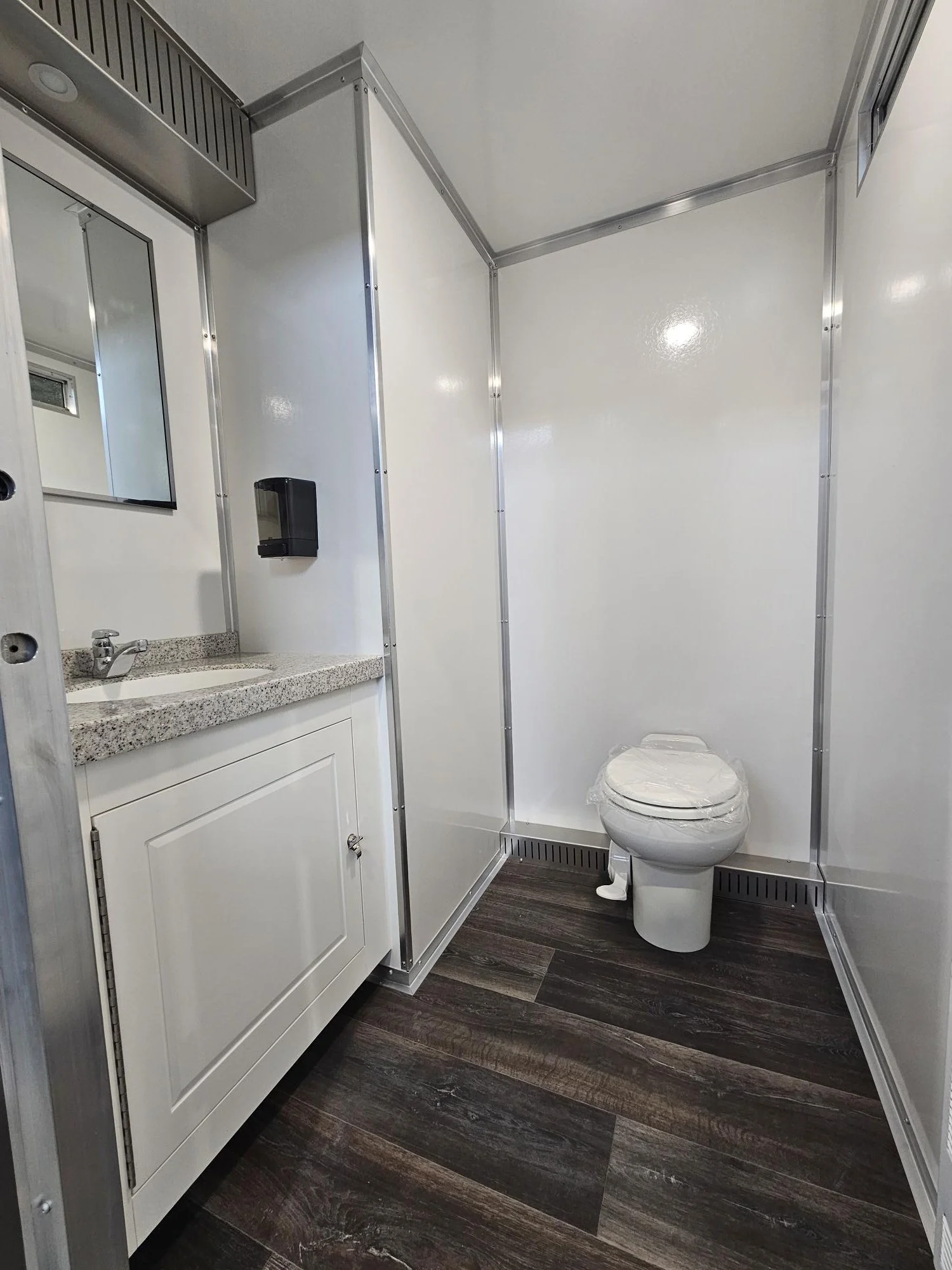 Rentable clean bathroom with white walls, dark wood flooring, a white sink vanity, a mirror, a black soap dispenser, and a toilet with a plastic cover.