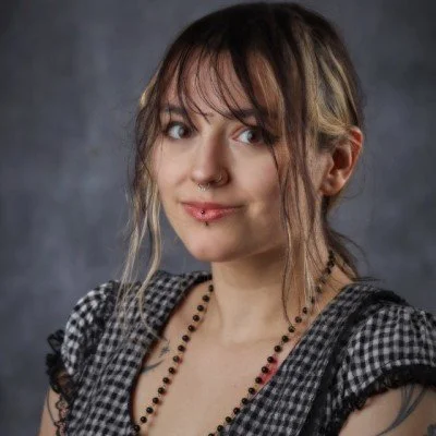 Young woman with light brown hair, wearing a black and white checkered top, a beaded necklace, and a nose and lip piercing, against a gray background.