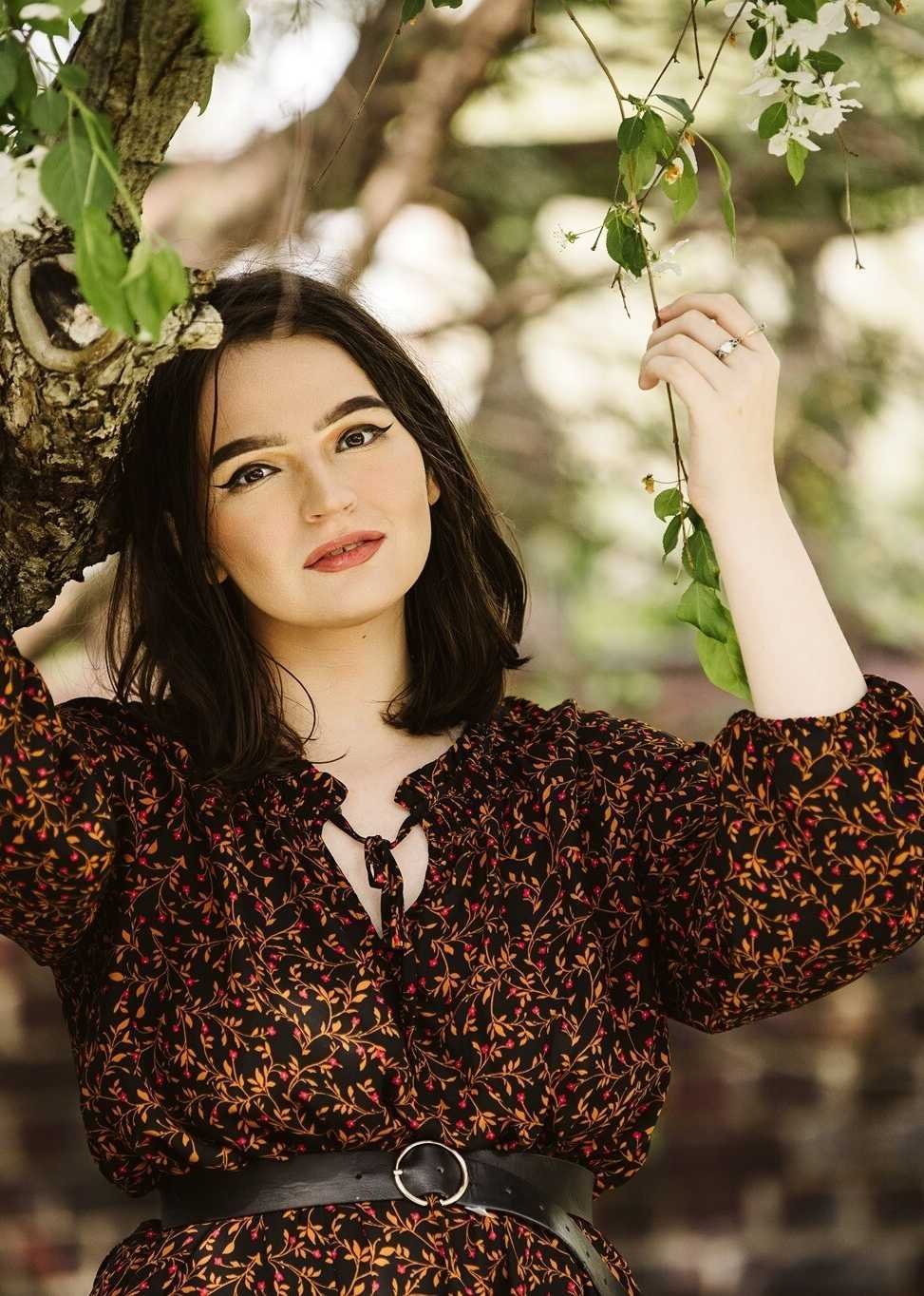 A young woman with black hair and makeup, wearing a black dress with a red and gold floral pattern, standing outdoors beside a tree and holding a branch with green leaves.