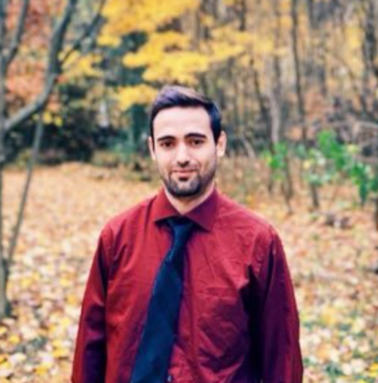 A man with dark hair and beard wearing a maroon shirt and dark tie, standing outdoors in a fall forest with yellow and orange leaves on the trees and ground.
