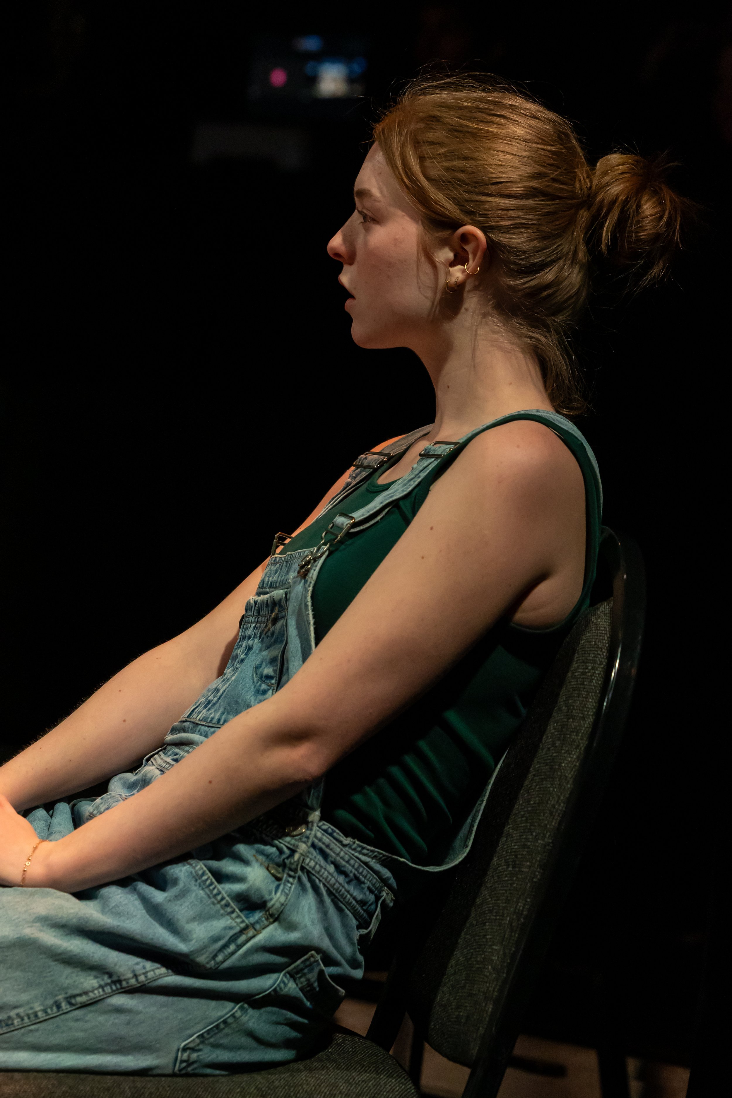 Side profile of a young woman with red hair tied in a bun, sitting on a chair, wearing a green tank top and denim overalls, against a dark background.