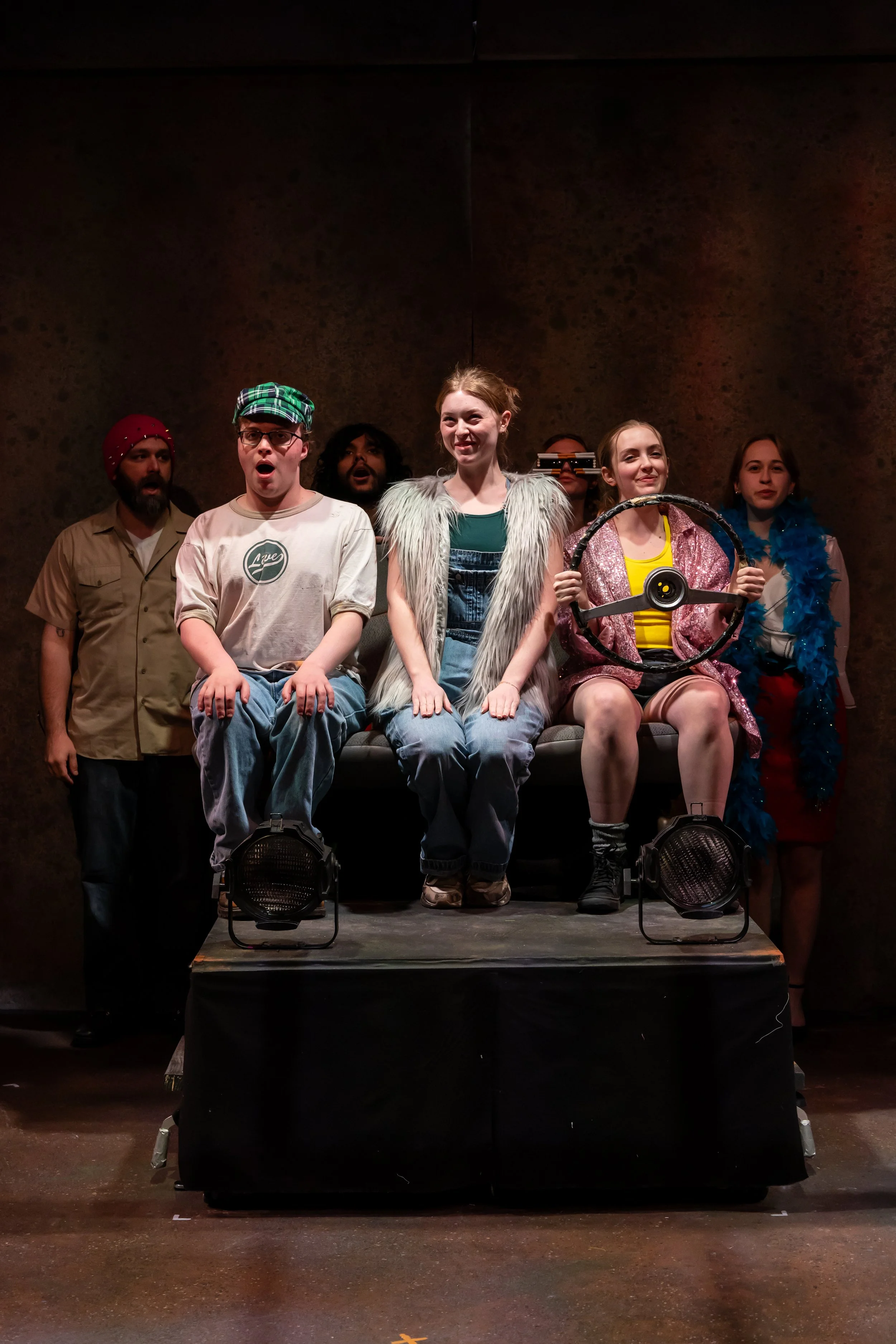 A theater cast performing on stage with six actors, some sitting on a bench and some standing behind, with one holding a steering wheel prop, wearing colorful costumes and expressing a variety of emotions.