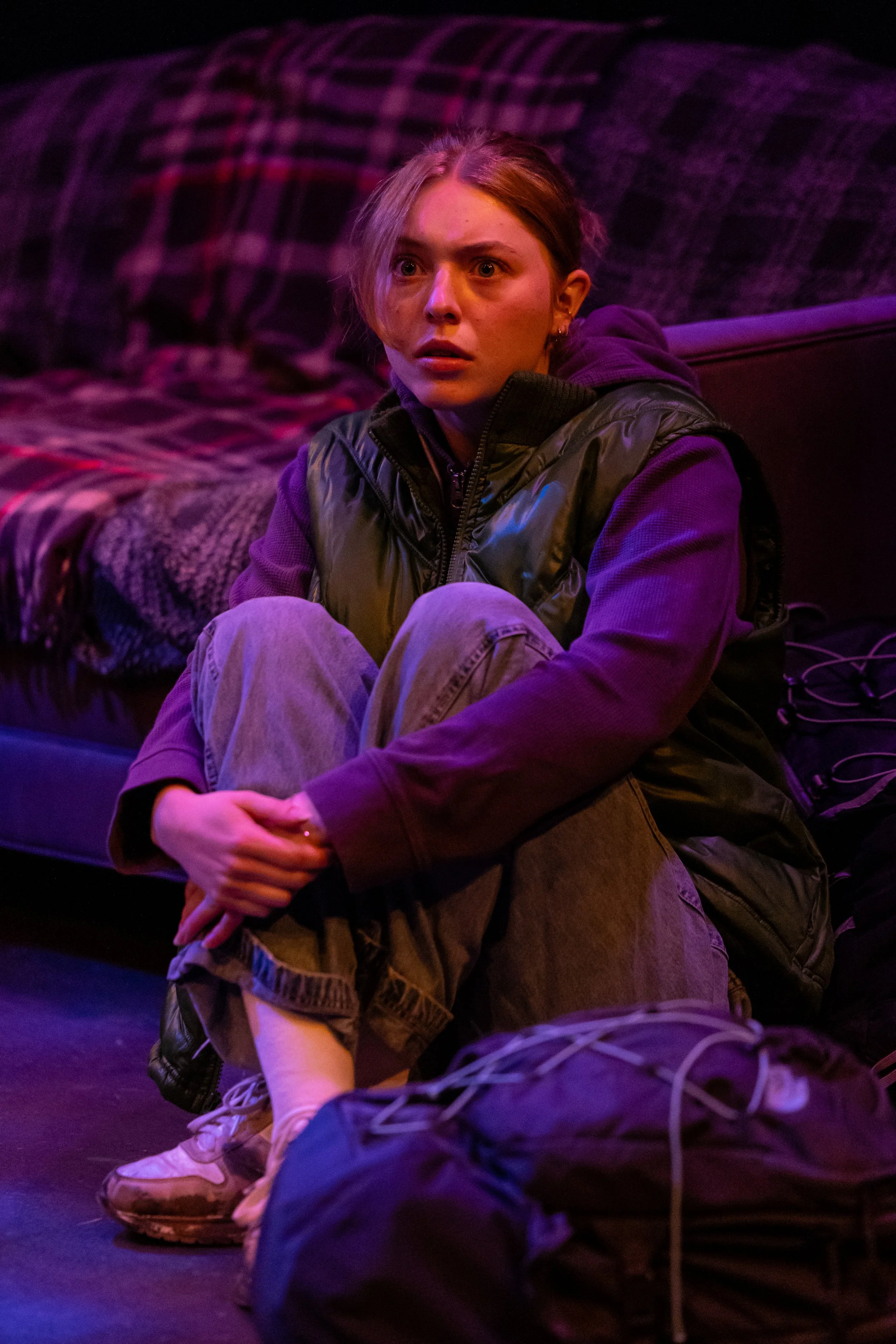 A young woman with a surprised or shocked expression, sitting on the floor with her knees drawn up, in a dimly lit room with purple hues, next to a bed with plaid blankets and a mess of cords and belongings.