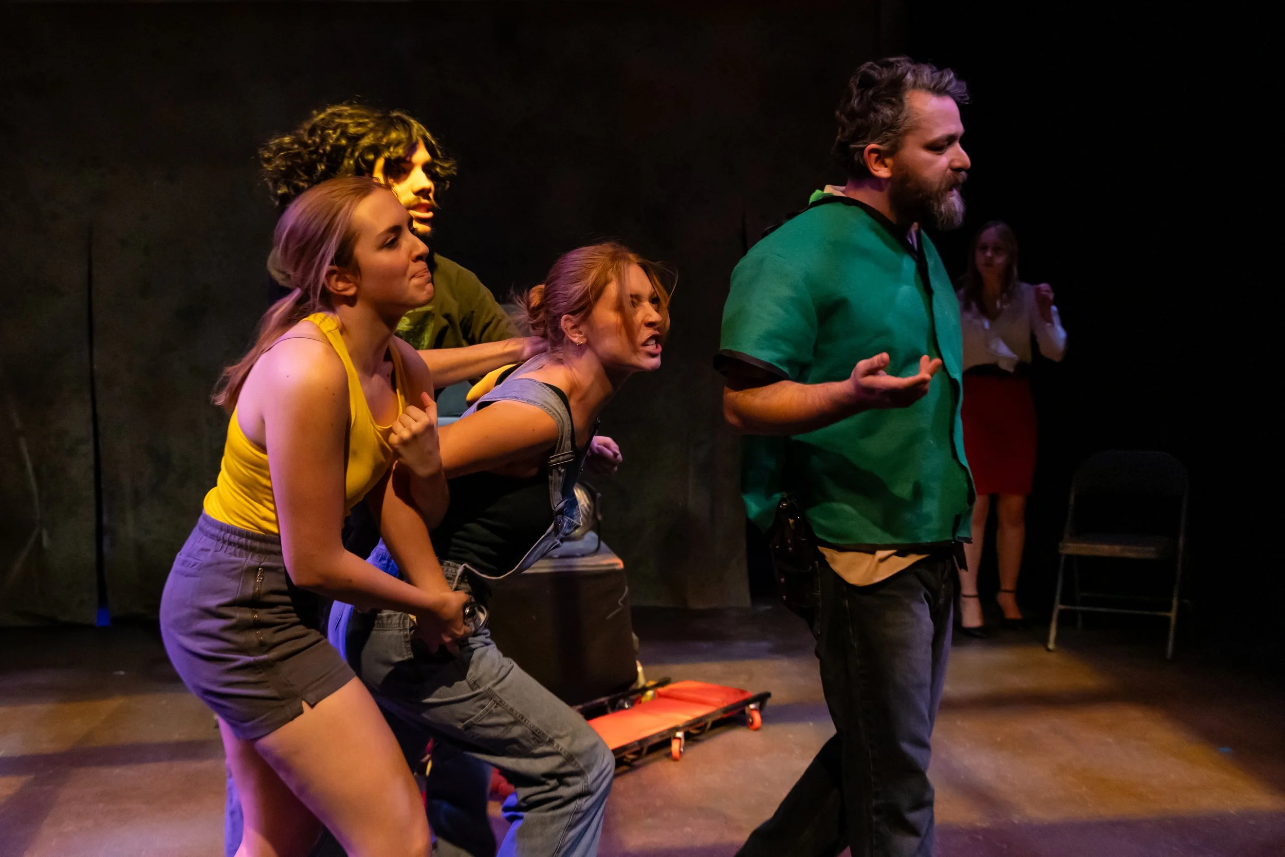 Five actors performing on stage with black background; three women and two men, some showing intense emotions, engaged in a theatrical scene.