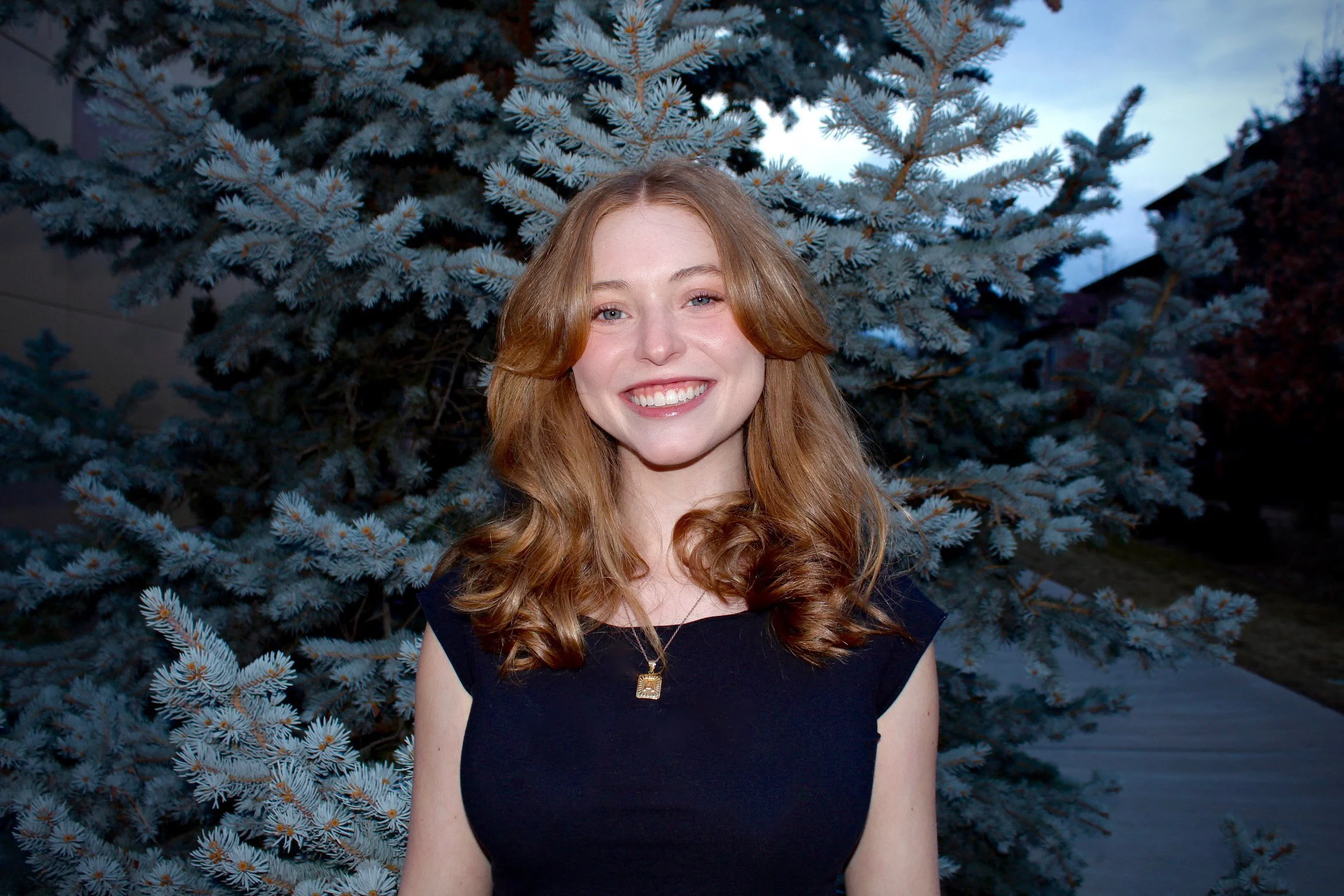 An image of Averie Dow, a young UX Professional with red hair wearing a black shirt in front of green trees. She is smiling.