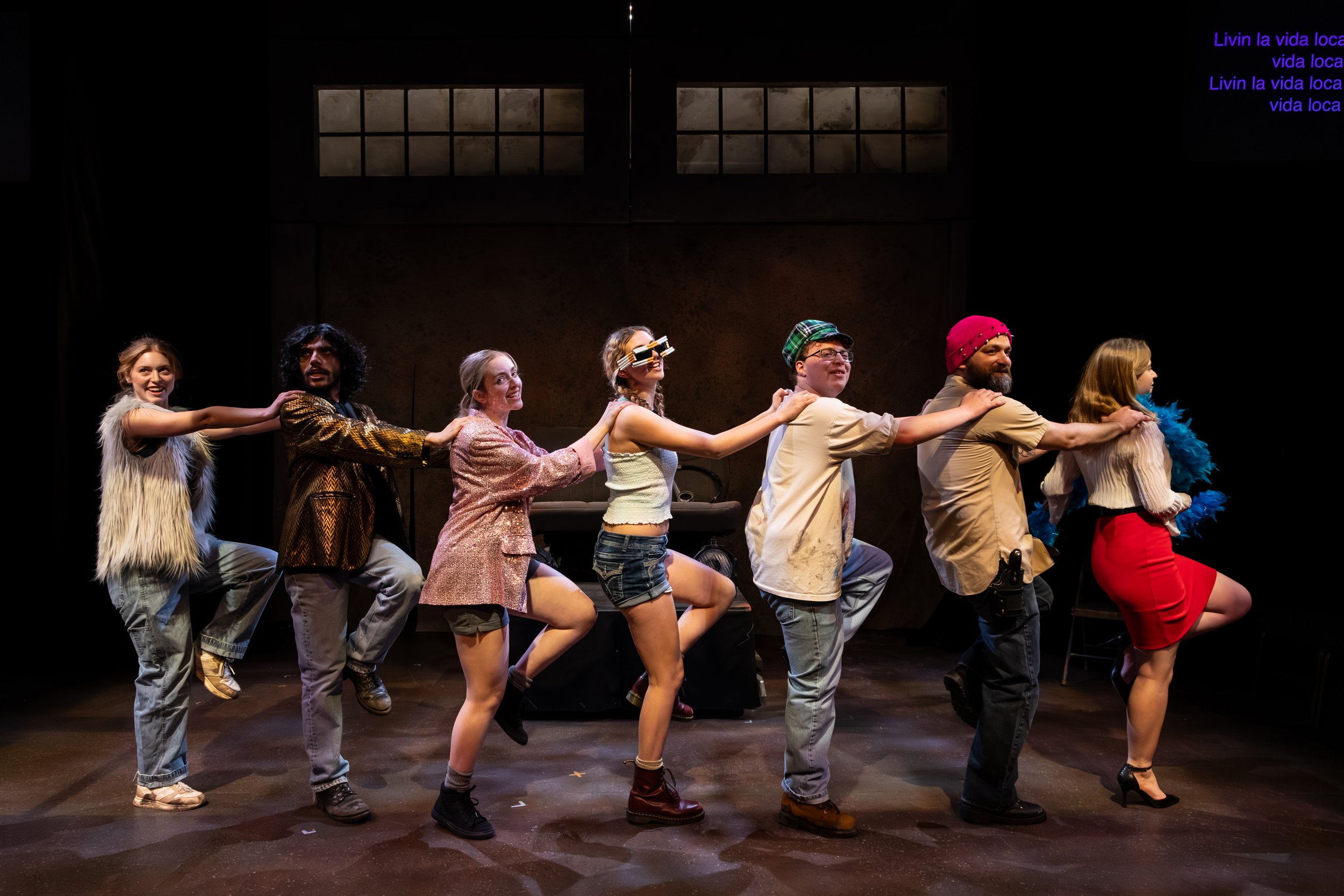 Group of diverse people participating in a theatrical performance, standing in a line with hands on each other's shoulders, on a stage with dark background.