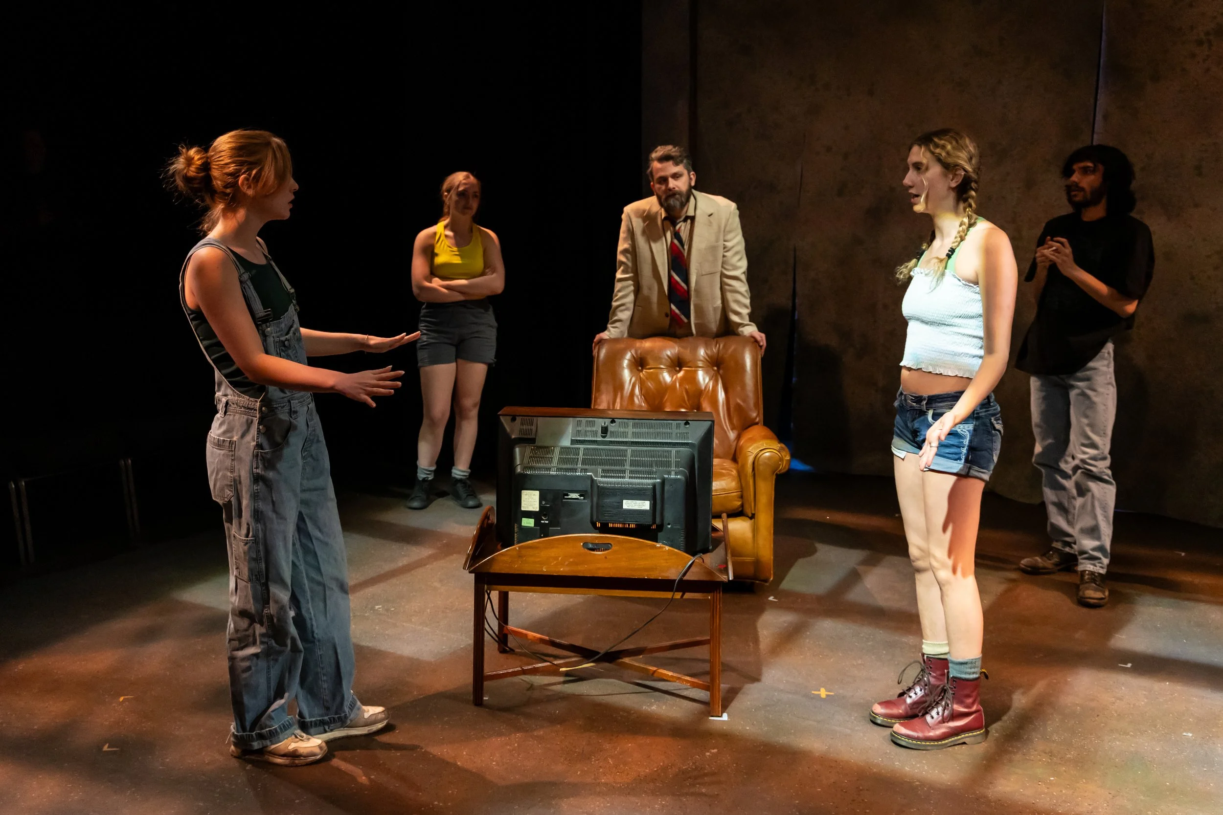 Four young women and one man standing on a stage, engaged in a conversation, with a TV on a table and a brown leather armchair behind them.