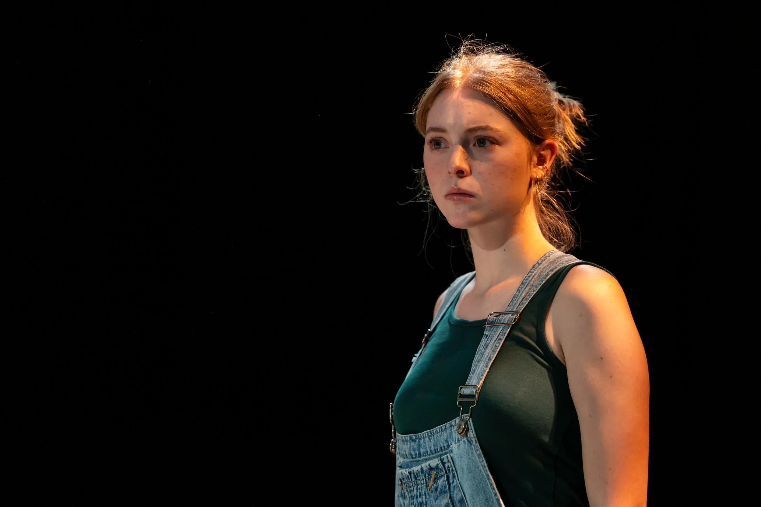A young woman with red hair and a serious expression stands against a black background. She is wearing a green tank top and denim overalls.