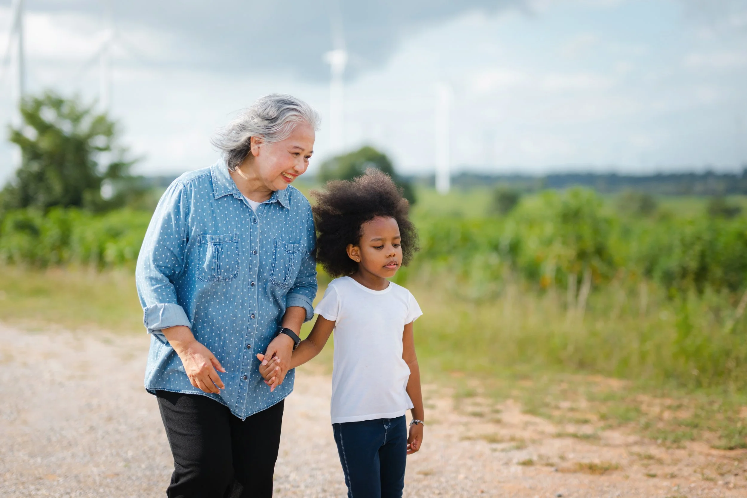 An elderly woman with gray hair and a young girl with curly hair hold hands and walk outdoors in a green field on a cloudy day.