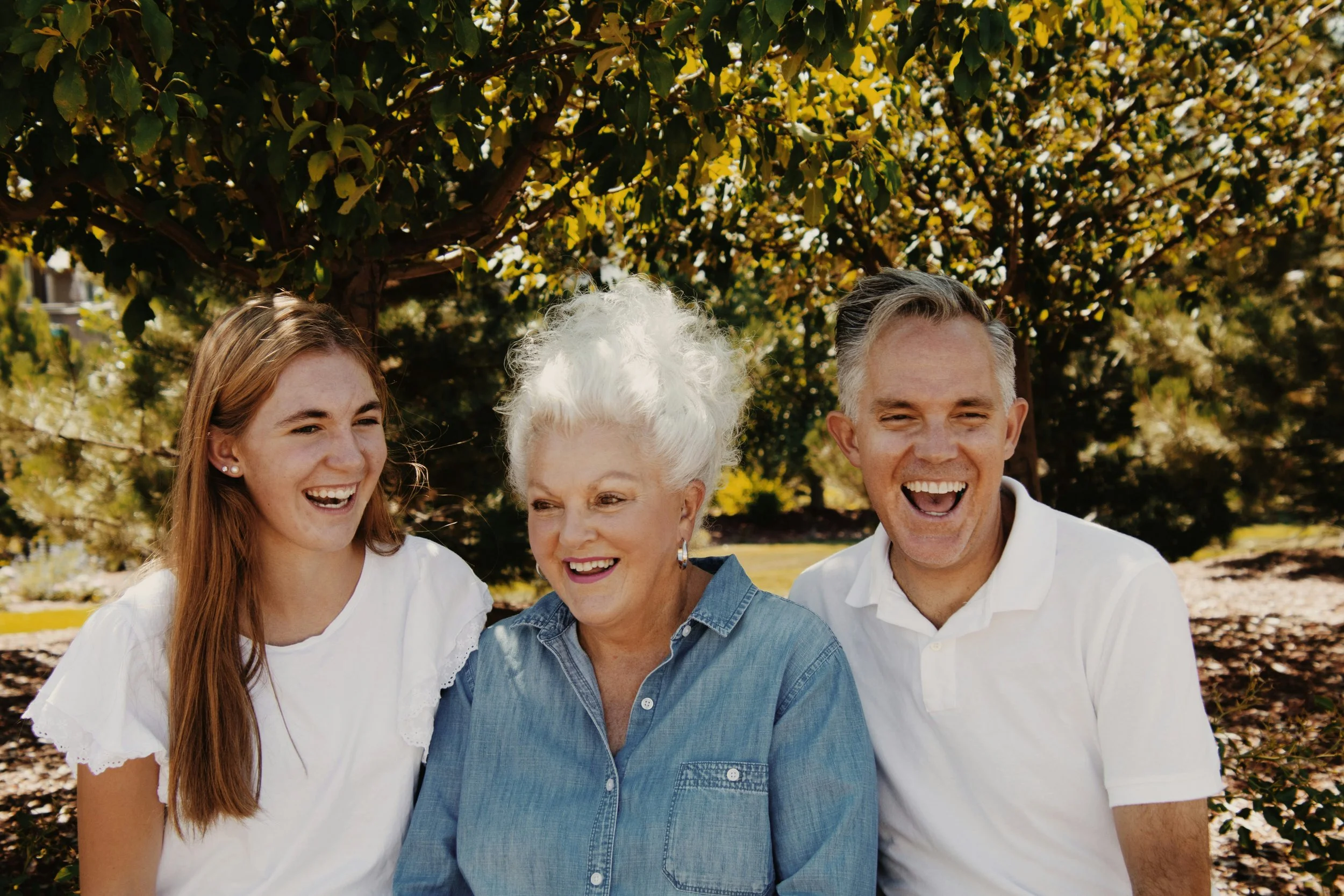 A group of four people, including a teenage girl, an elderly woman, and two adult men, smiling outdoors in front of trees with yellow and green leaves.