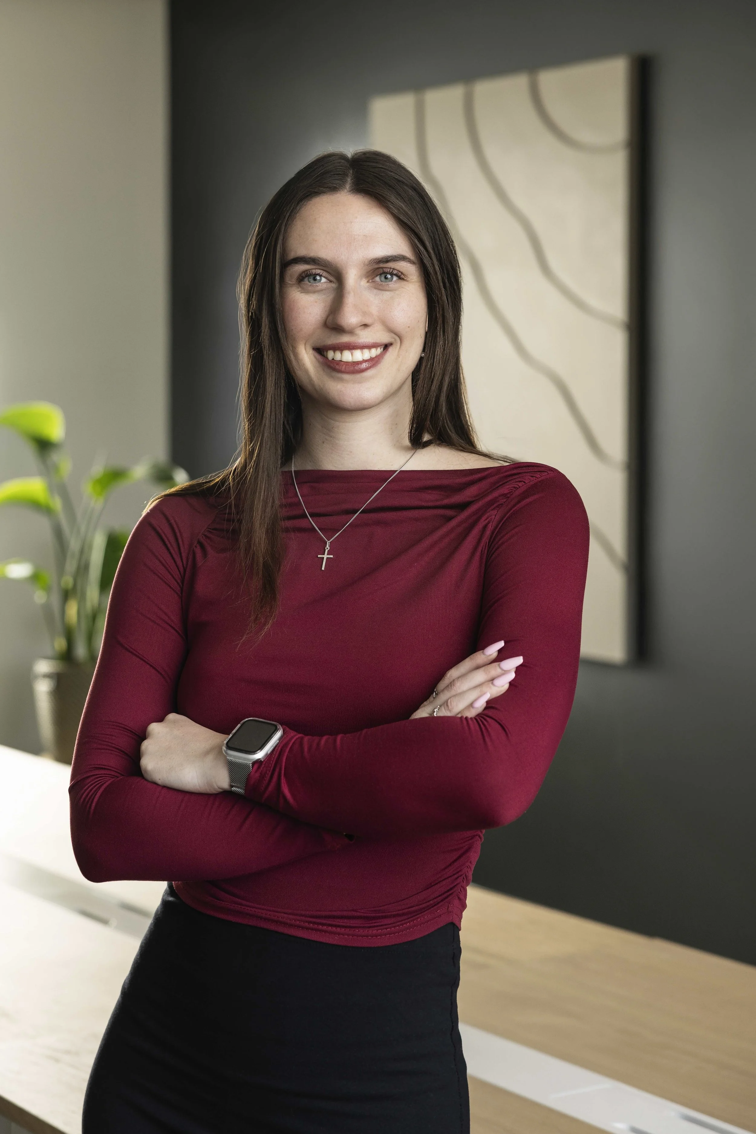 A woman with long brown hair and blue eyes smiling, standing with arms crossed in a modern office with a plant and abstract artwork in the background.