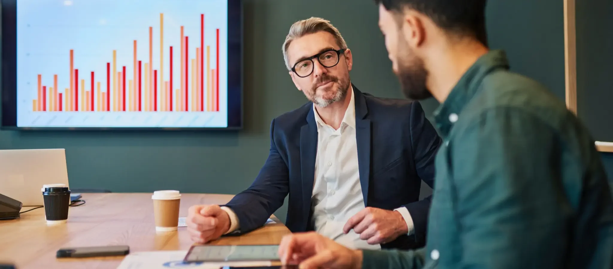 Two men in a meeting room having a discussion, with a bar chart displayed on a screen behind them.
