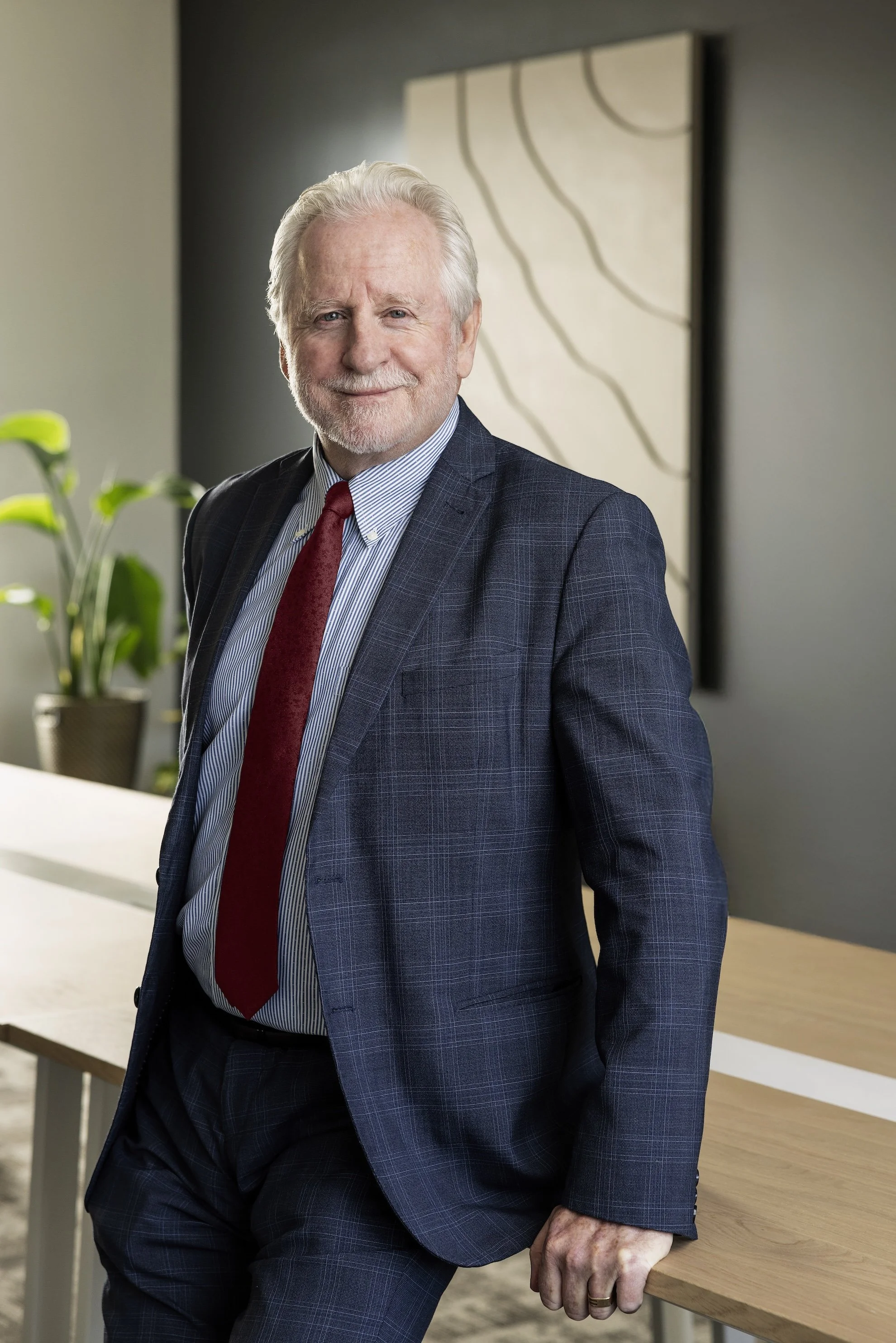 An elderly man dressed in a dark blue plaid suit with a red tie stands beside a wooden table in a modern office setting, smiling at the camera.