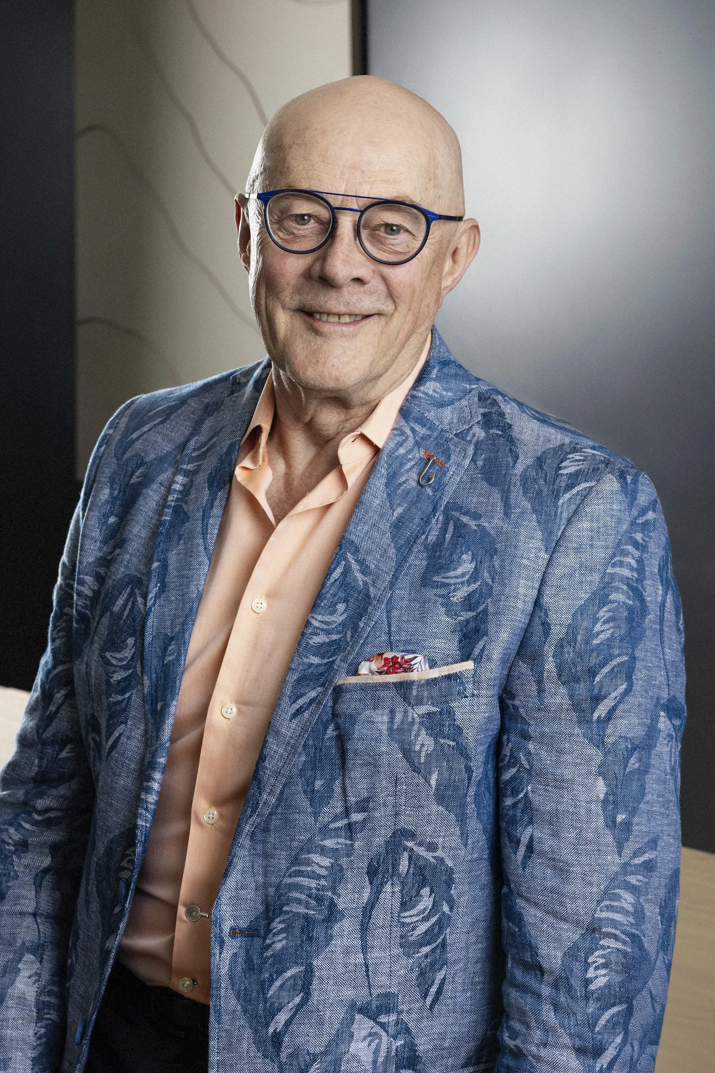 An older man with glasses and a balding head, smiling, wearing a patterned blue blazer, light orange shirt, and a pocket square, standing indoors with a neutral background.