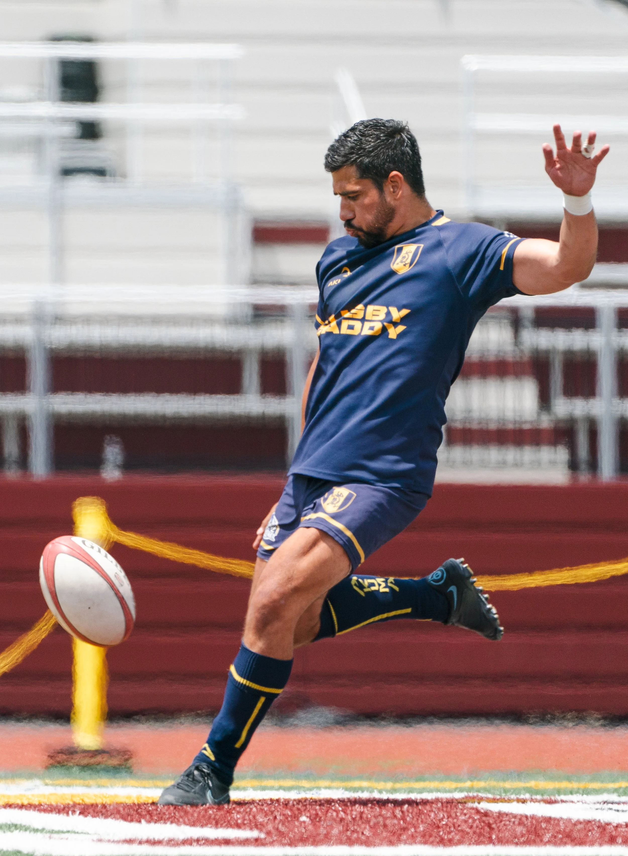 A Gotham Knights player in navy and gold mid-kick, sending the ball downfield during a match.