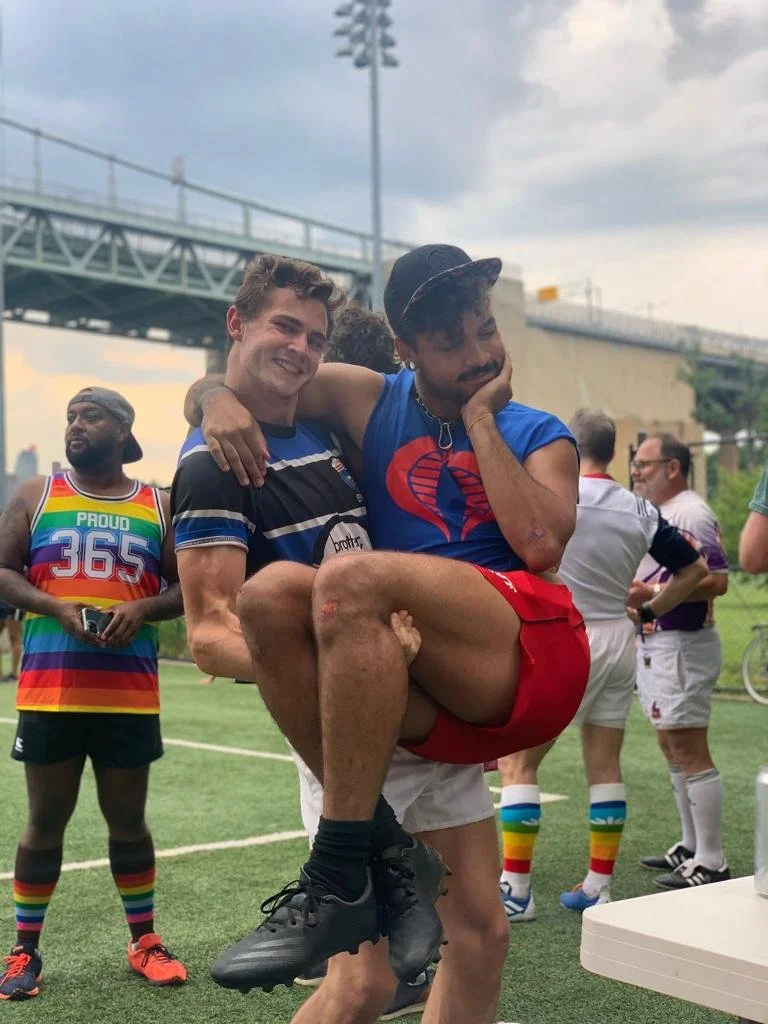Two Gotham Knights players clowning around after practice, one carrying the other on his back, with teammates in rainbow jerseys behind them.