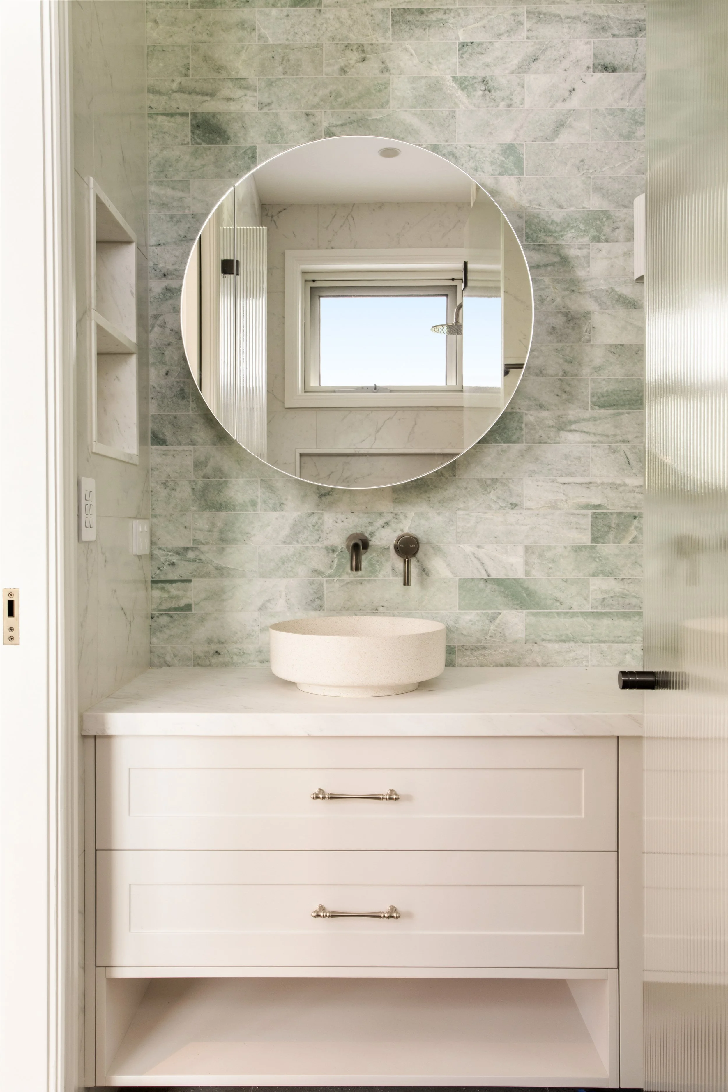 Bathroom vanity with a round mirror, marble backsplash, white cabinetry, vessel sink, wall-mounted faucet, window reflected in mirror, and partial view of shower door.