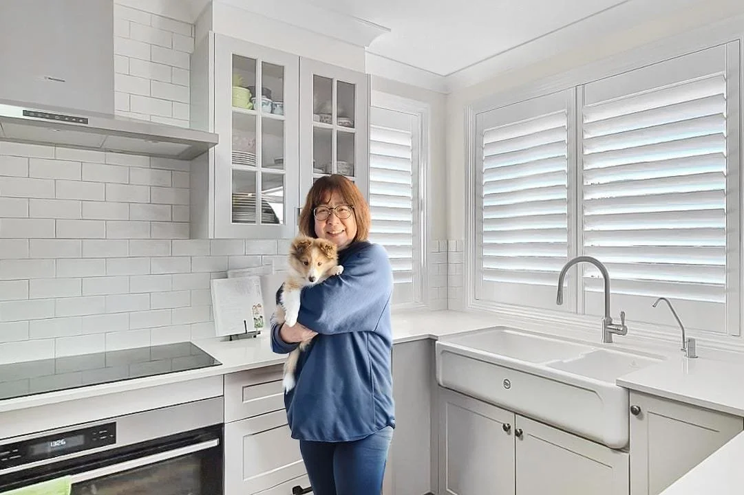 A woman holding a small dog in a white kitchen with shaker cabinets, a farmhouse sink, window blinds, and white tiled backsplash.