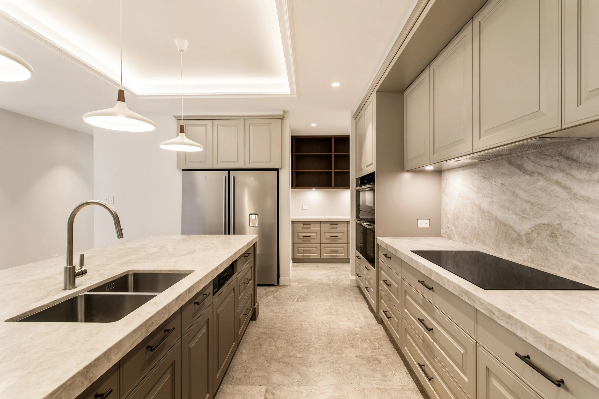 Modern kitchen with beige cabinets, marble countertops, and built-in appliances, including a double oven and cooktop, illuminated by pendant lights.