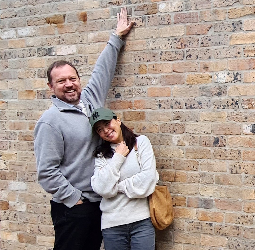 A man and woman with light skin posing against a brick wall. The man, wearing a gray sweatshirt, is smiling and raising one hand, touching the wall. The woman, wearing a beige sweater and a green baseball cap, is smiling, with her tongue out, and her hand near her chin. She carries a tan shoulder bag.
