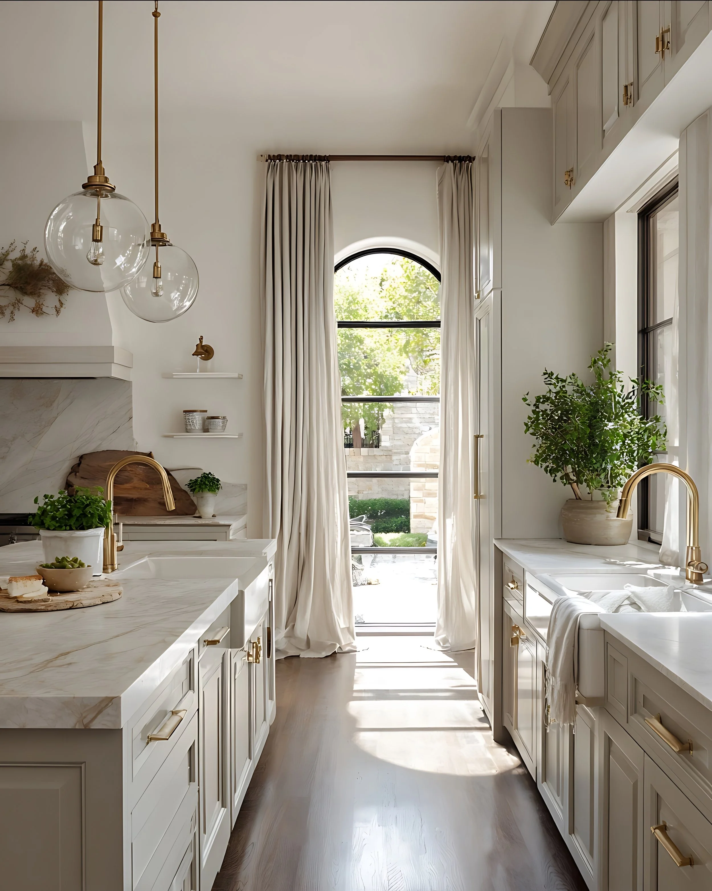 Bright modern kitchen with white cabinets, gold fixtures, marble countertops, and large windows with curtains, overlooking a garden.