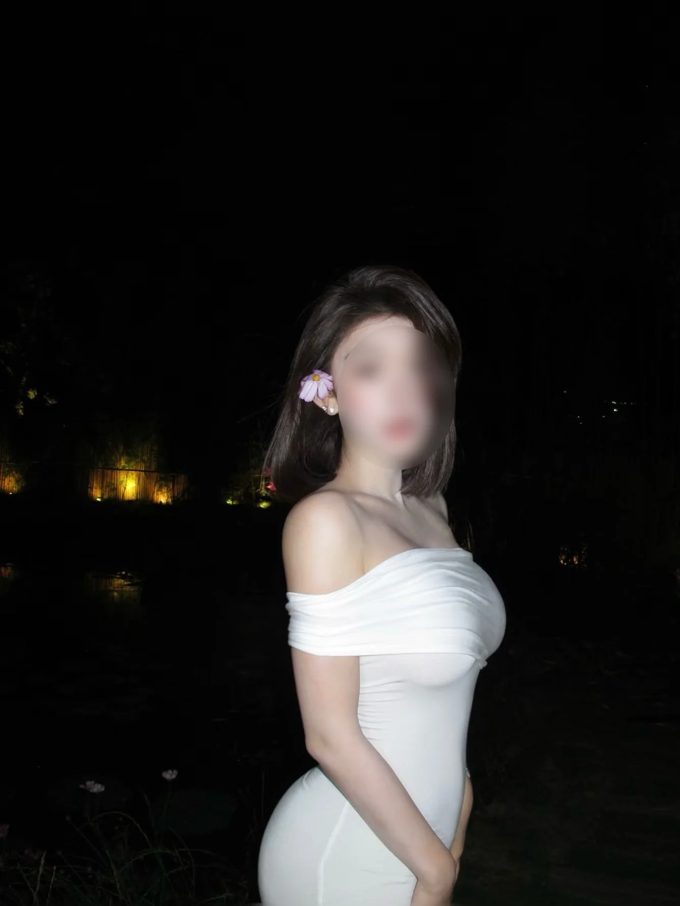 A woman in an off-shoulder white dress with a flower in her hair standing outdoors at night.