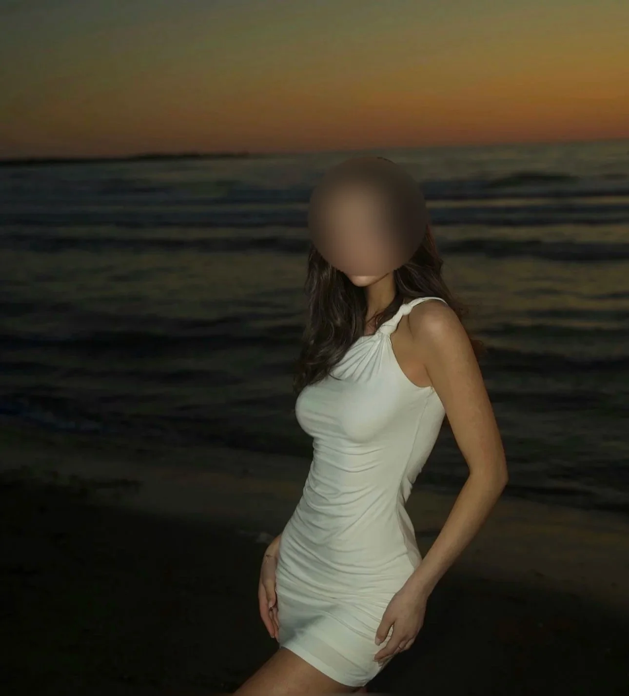 A woman in a white dress standing on the beach at sunset with the ocean in the background.