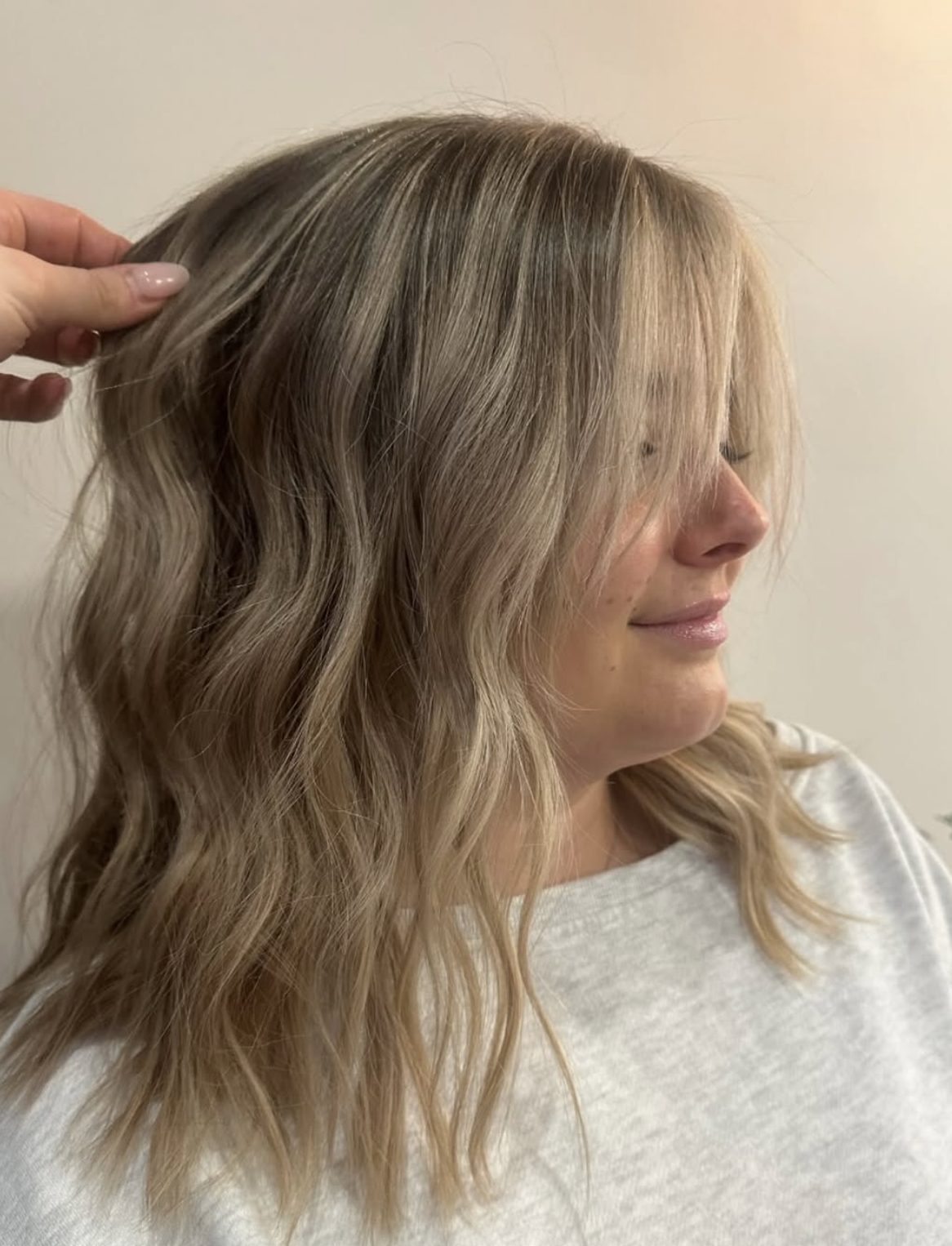 A woman with shoulder-length blonde, wavy hair, smiling gently, with someone touching her hair.