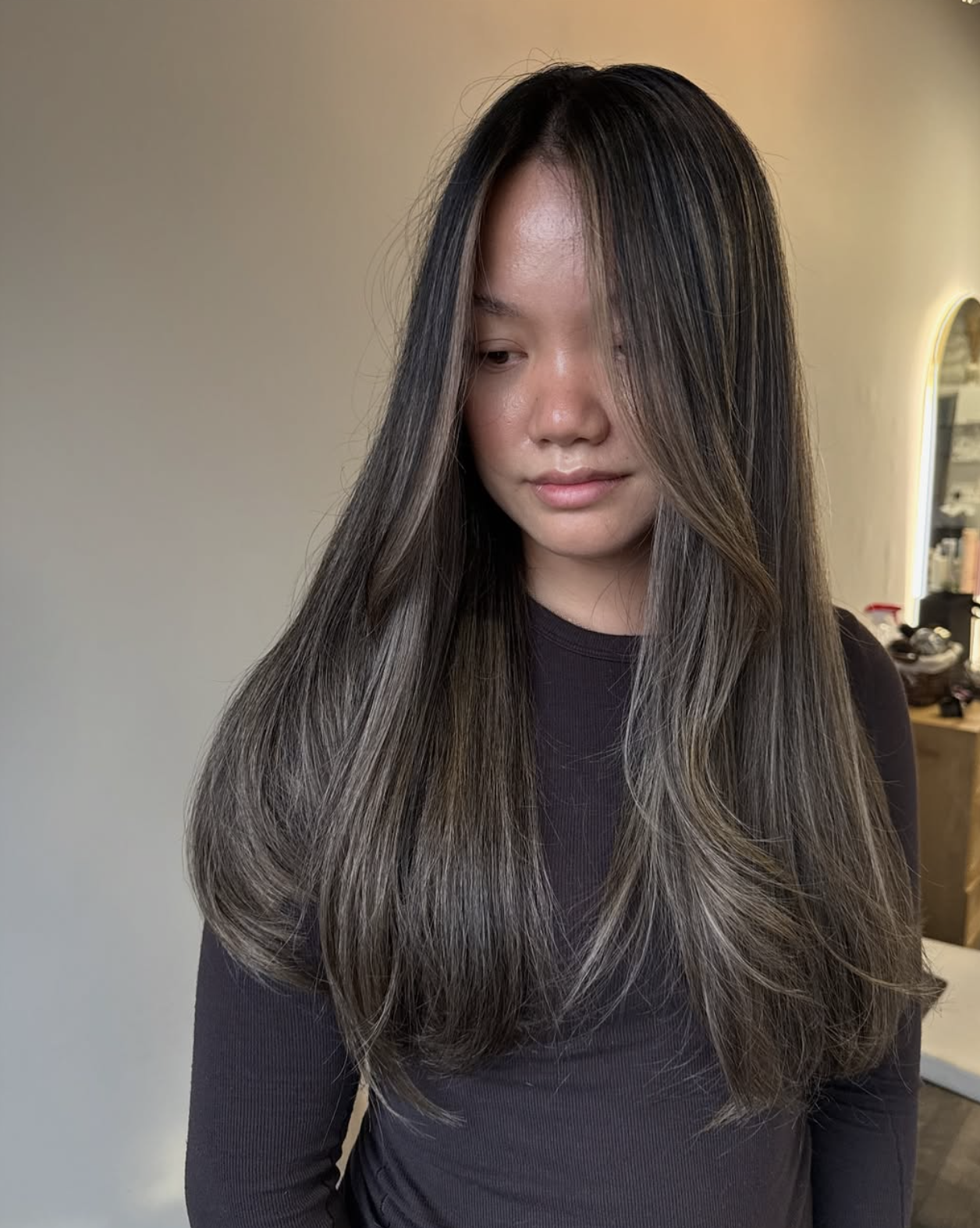 A woman with long dark hair with lighter streaks, wearing a dark long-sleeve shirt, looking down with a neutral expression in an indoor setting.