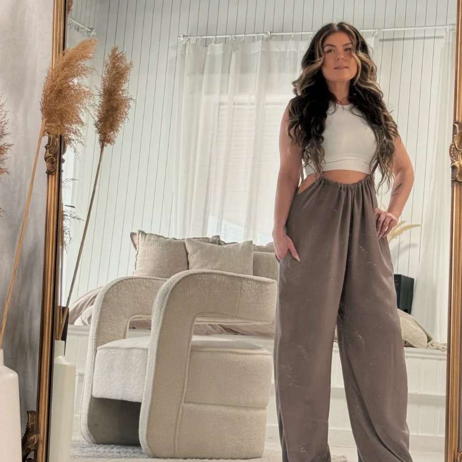 A woman takes a mirror selfie in a cozy, light-colored living room. She is wearing a white cropped sleeveless top and loose brown pants. The room features a beige armchair, beige pillows on a sofa, and decorative dried plants.