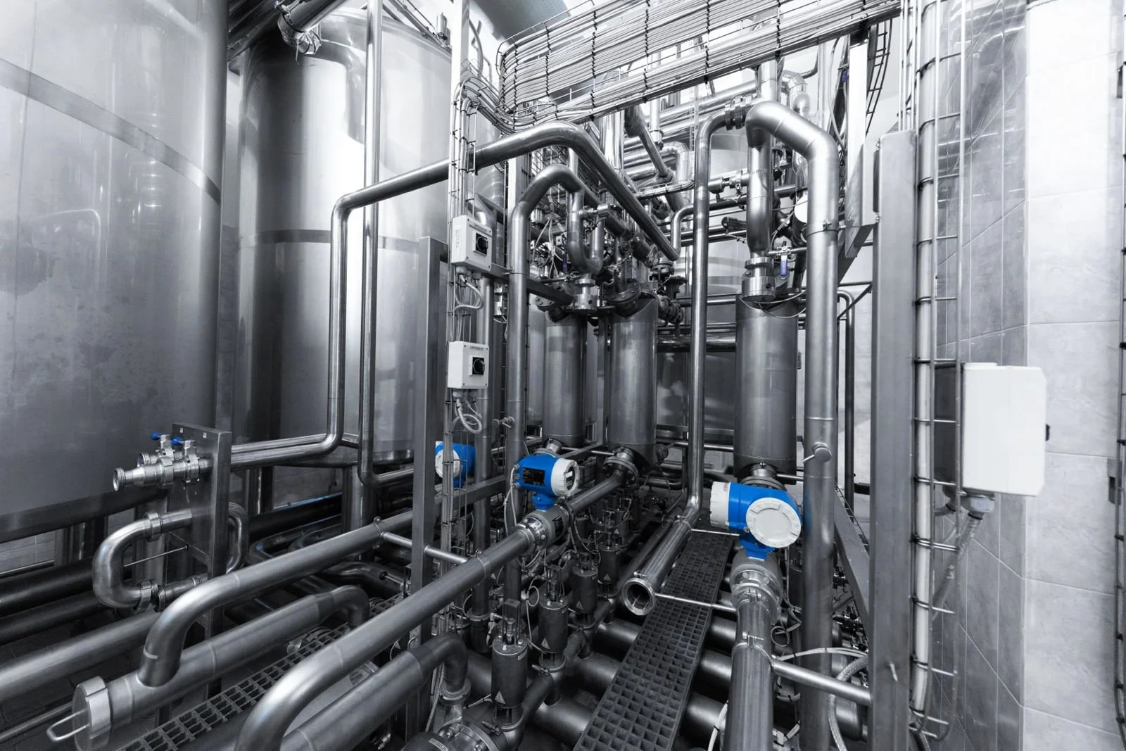 Industrial process room with stainless steel pipes, tanks, and blue valves.