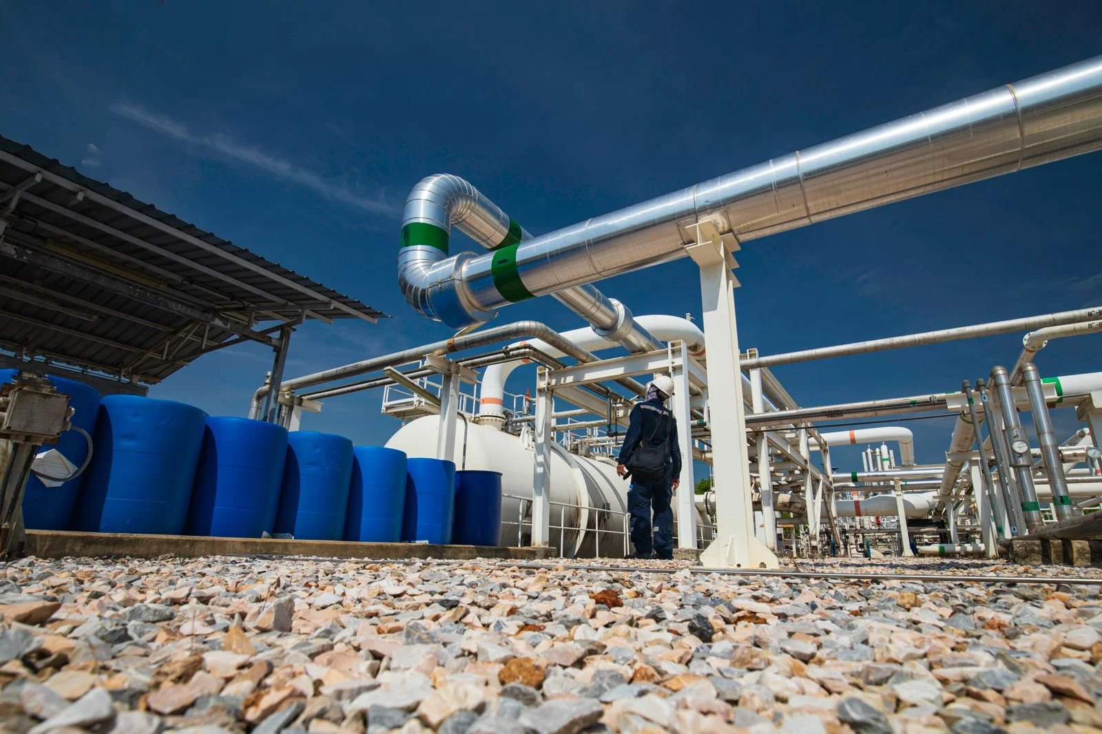 An industrial facility with pipelines and blue tanks, a person wearing safety gear walking among equipment, under a blue sky.