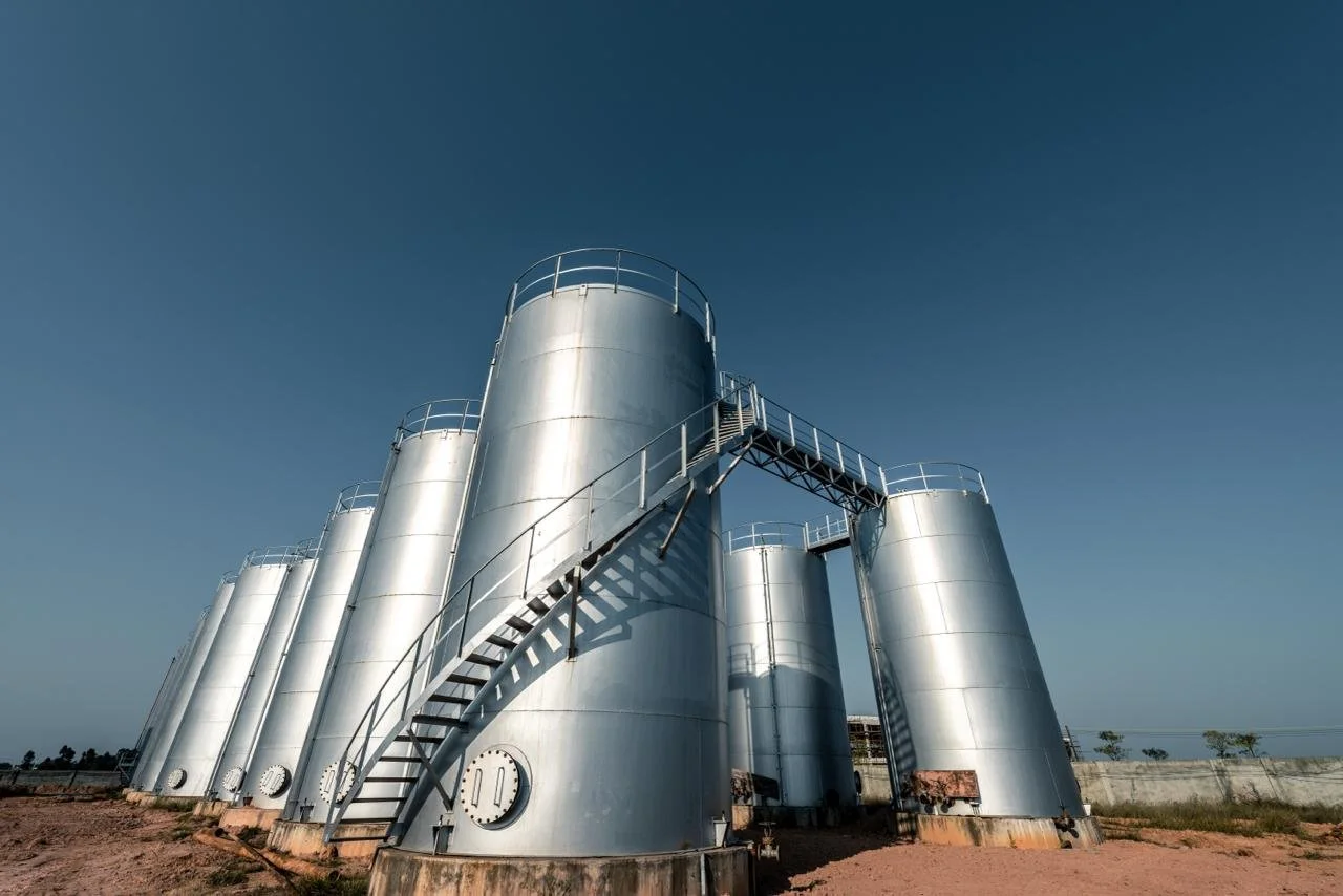 Large metallic industrial storage tanks connected by stairs and walkways, set in an open area under a clear blue sky.