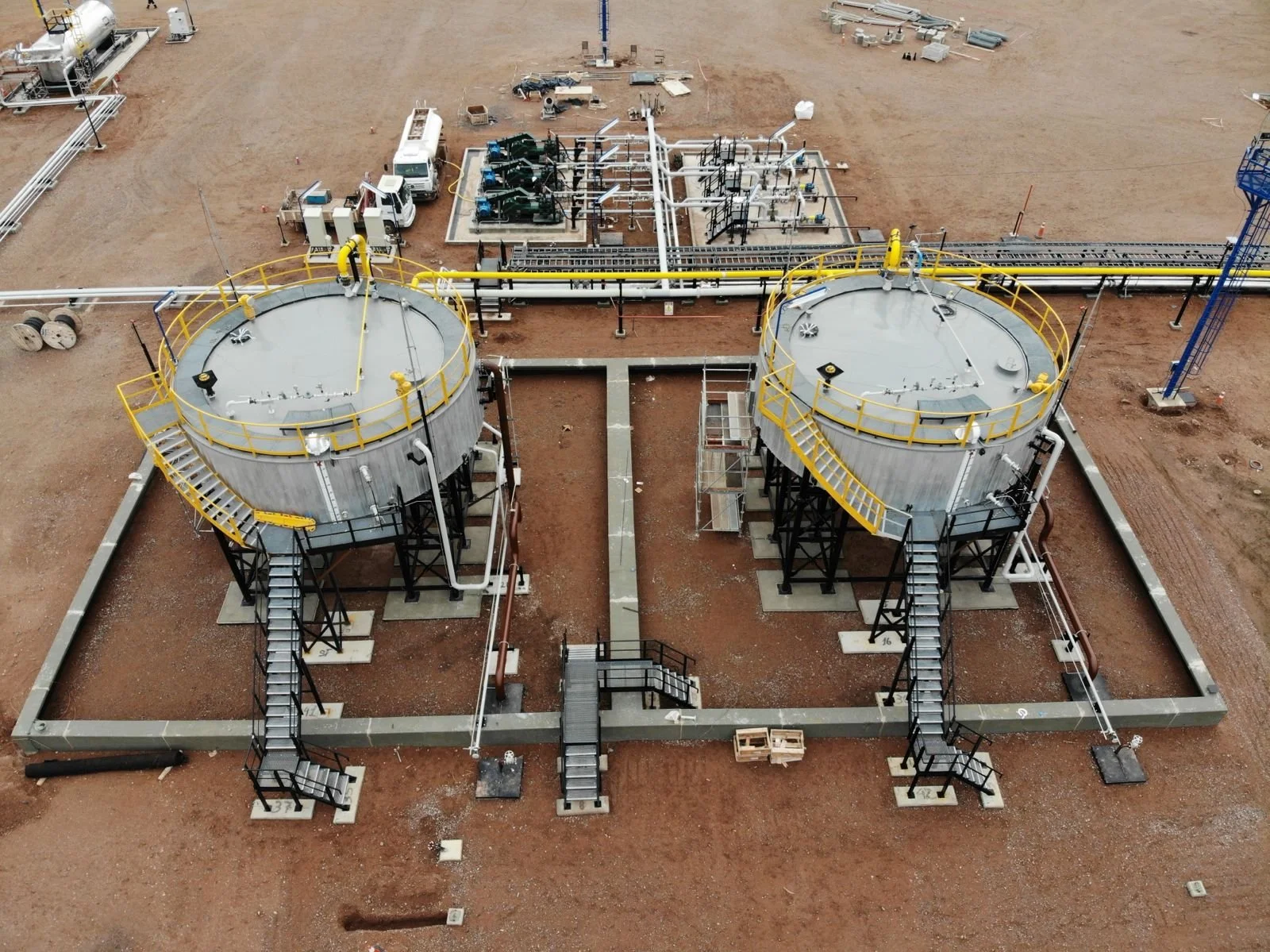 Two large industrial storage tanks on a construction site with stairs and safety railings, connected by pipelines, surrounded by dirt ground.