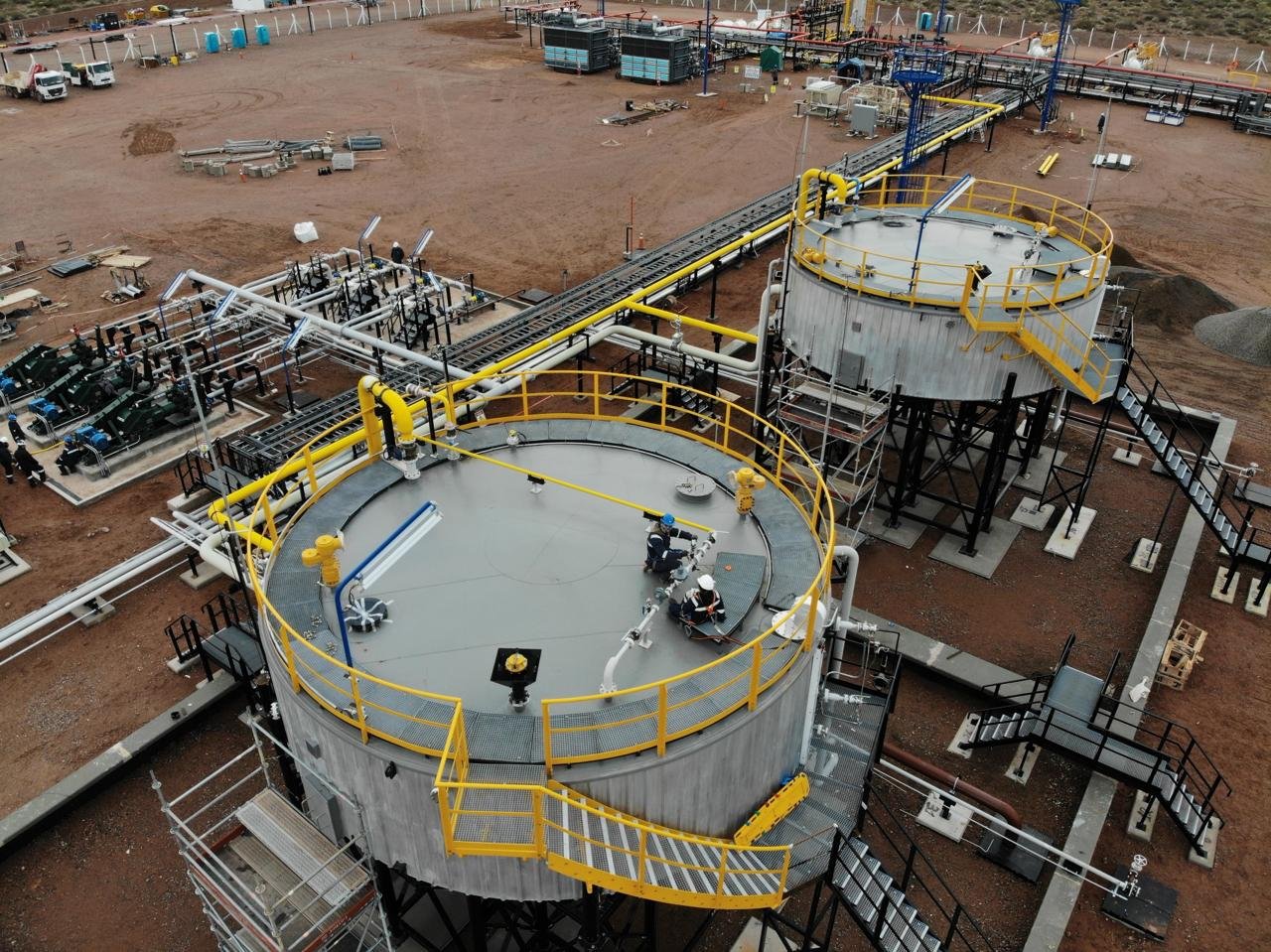 Two large metal industrial tanks with yellow railings, pipes, and stairs in an industrial facility. Workers in safety gear are working on top of the tanks, with machinery and equipment in the background.