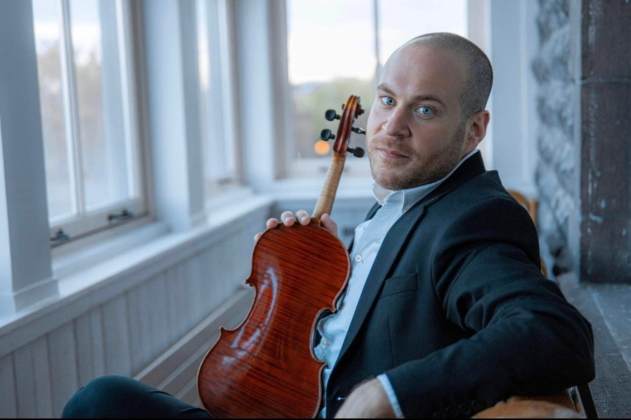Man in a black blazer holding a guitar on his shoulder indoors near a large window.