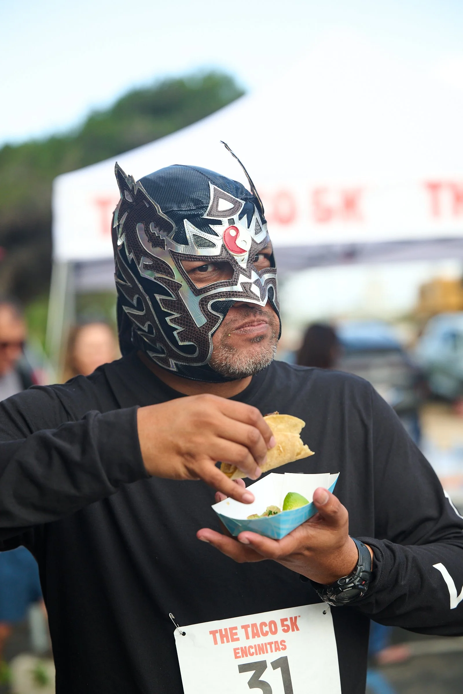 A man wearing a decorative lucha libre mask is holding a taco in one hand and a bowl of chips and lime in the other. He is participating in a race called 'The Taco 5K' based on his bib number.
