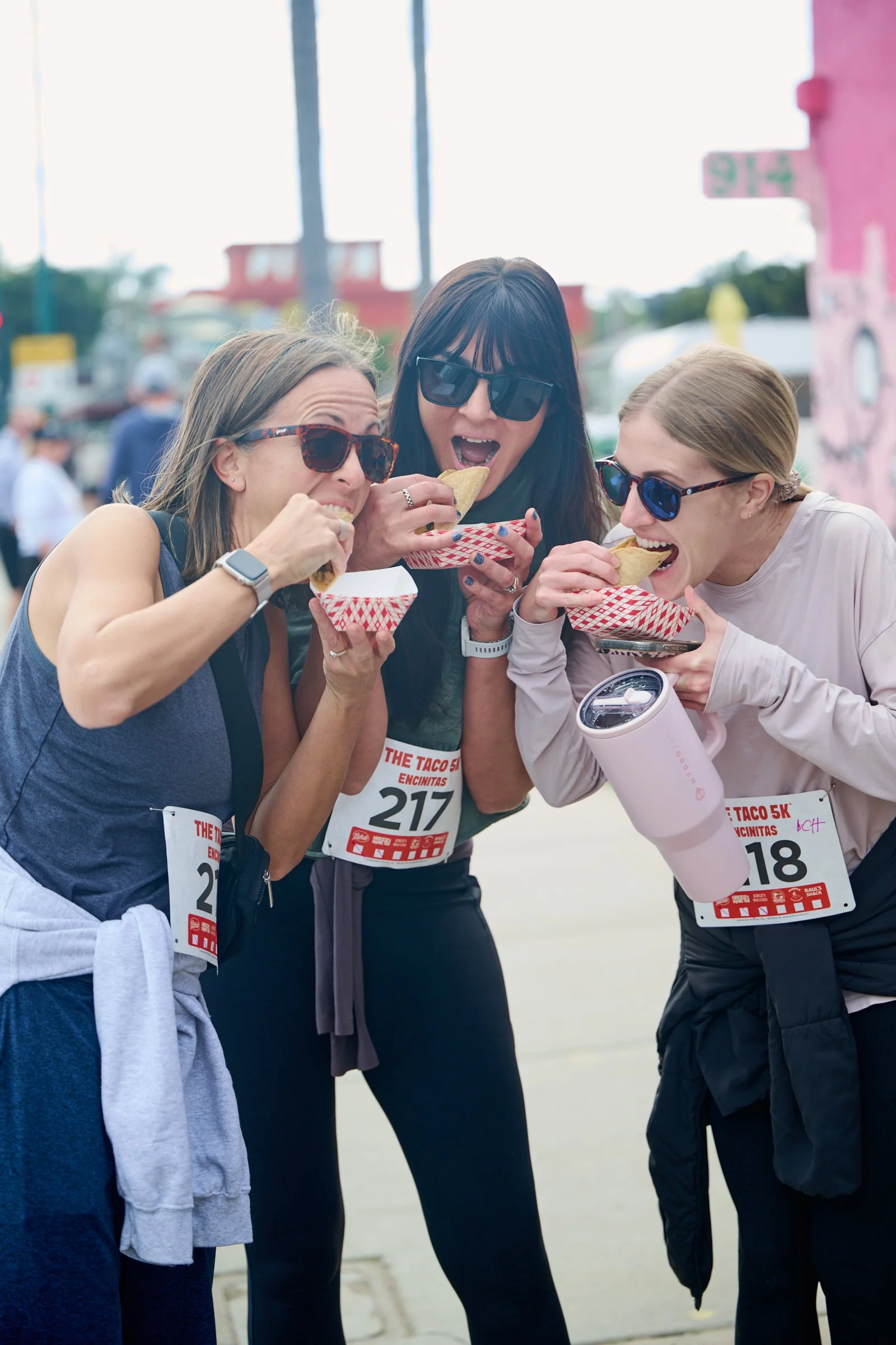 Three women wearing sunglasses and race bibs eating tacos at an outdoor event.