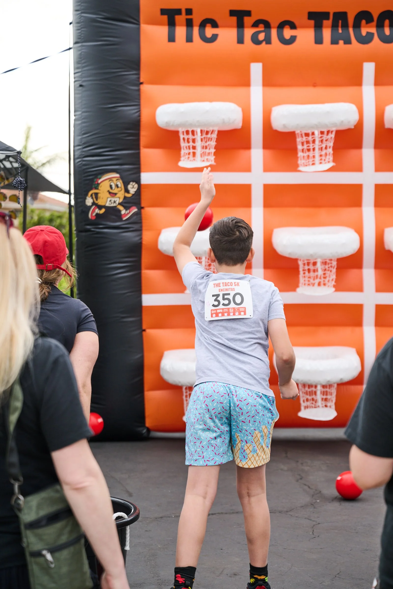 A young boy playing an outdoor basketball game where he is aiming to throw a red ball into a basketball hoop with a foam cushion top, set up on an orange and black inflatable structure.