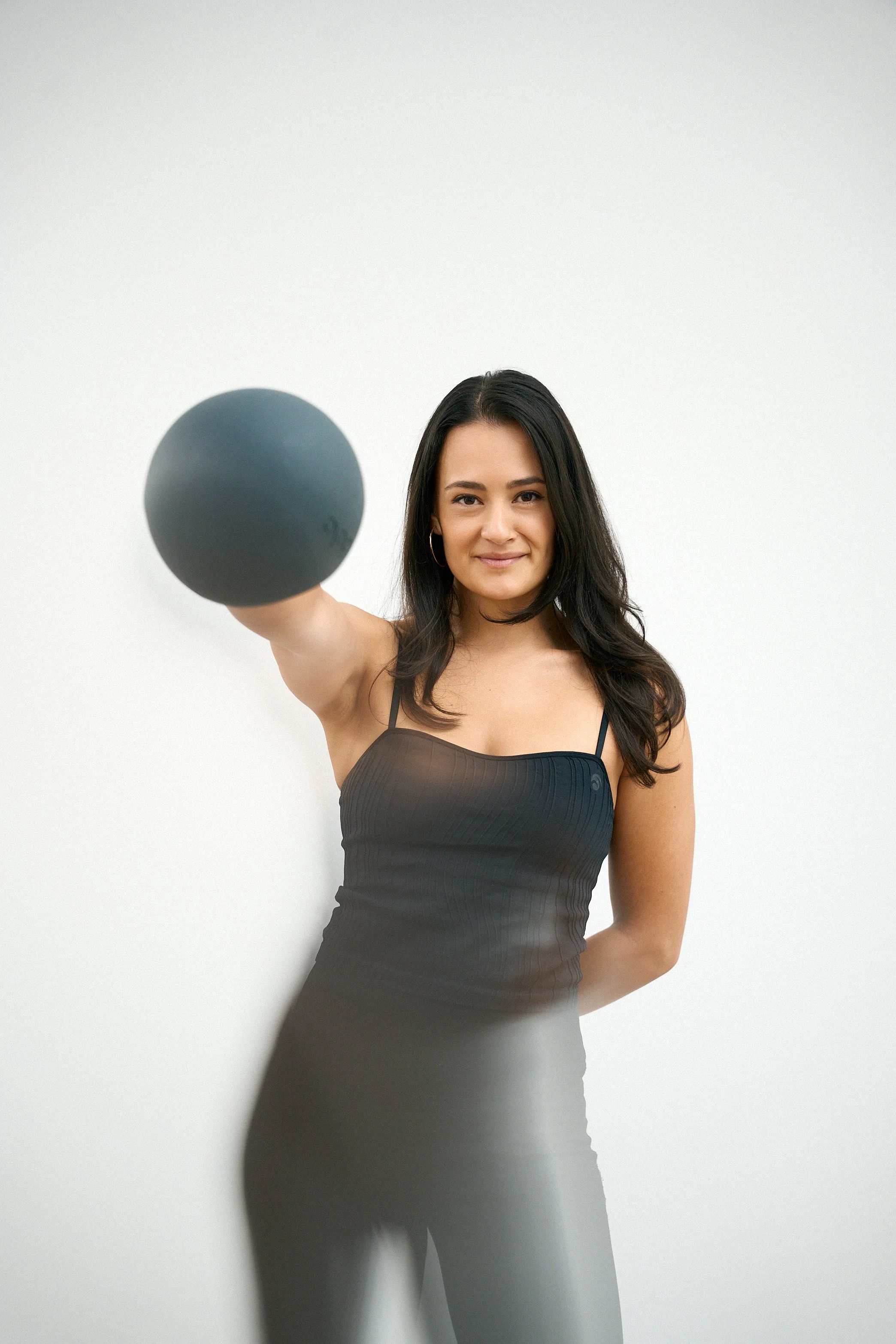 Junge Frau mit schwarzen Haaren und dunkler Kleidung hält einen schwarzen Ball in die Kamera, vor weißem Hintergrund.