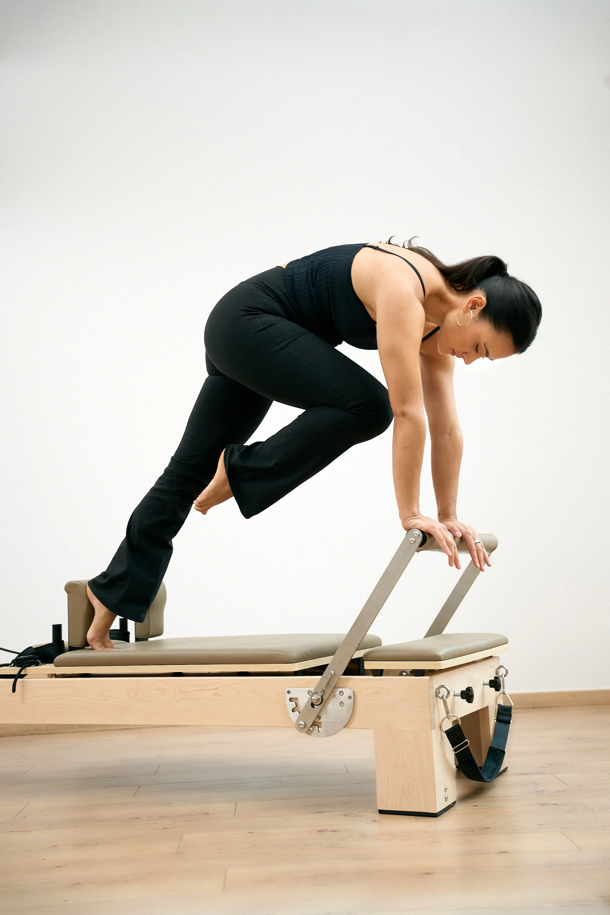 Frau macht Yoga auf einem Pilates-Reformer in einem Raum mit Holzfußboden und weißer Wand.