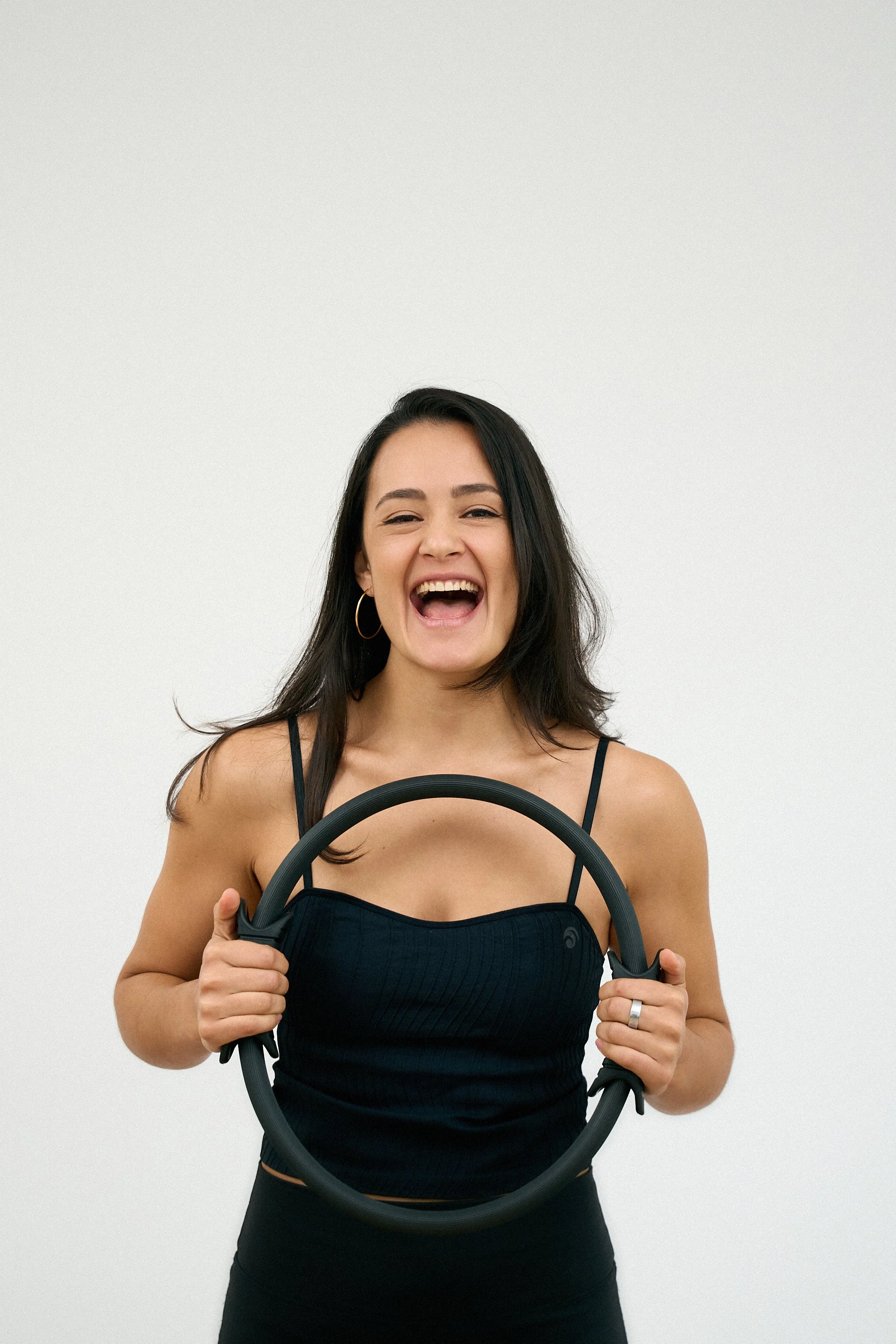 Lächelnde junge Frau mit dunklen Haaren trägt schwarzes Top und hält ein Trainingsband vor weißem Hintergrund.