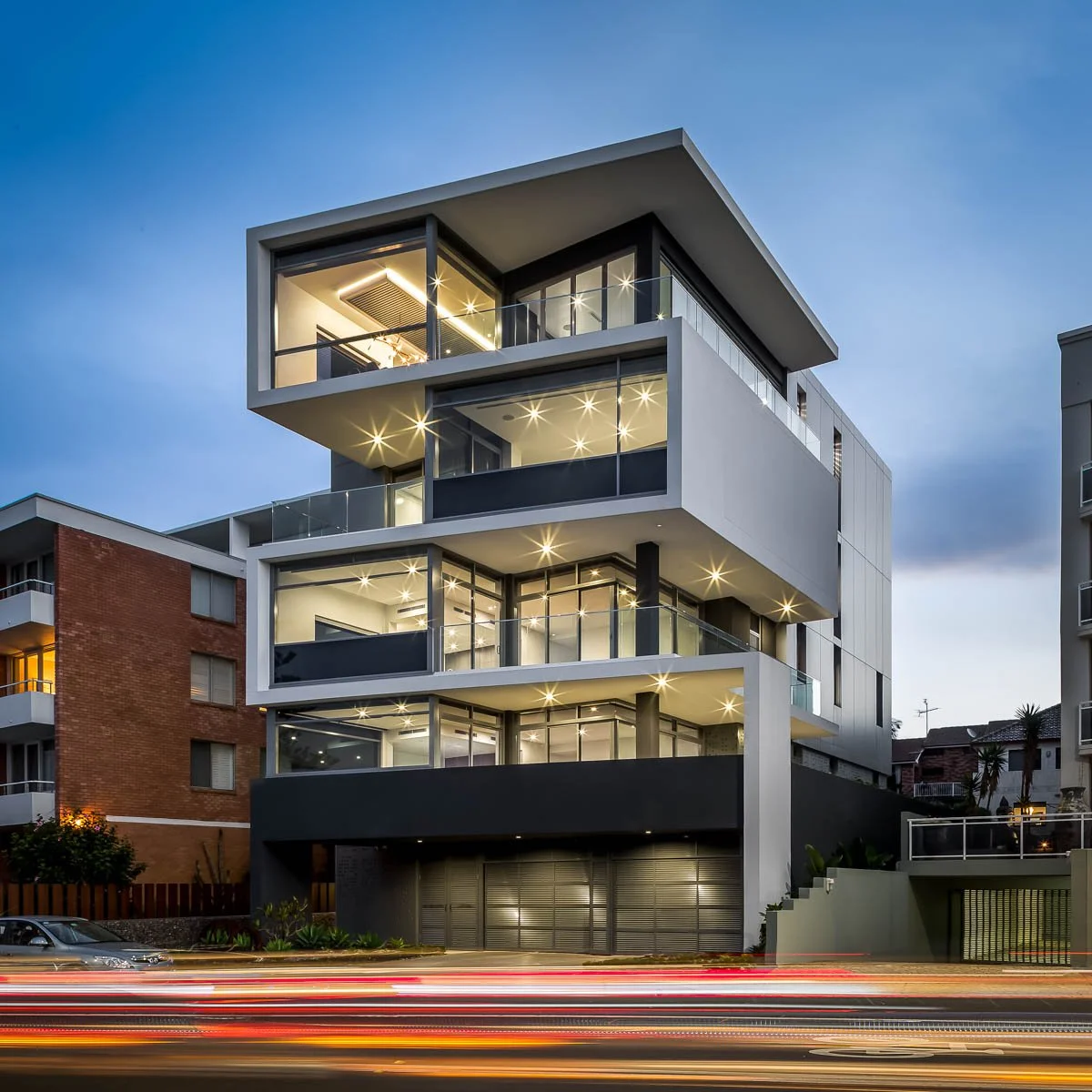 Modern multi-story apartment building with multiple lit balconies and glass railings during dusk, neighboring buildings visible.