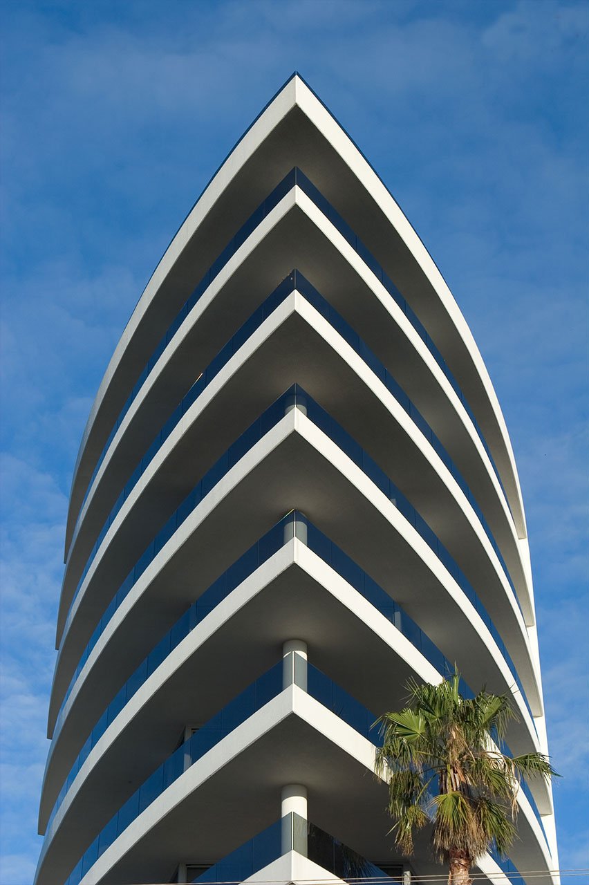 Tall modern building with curved white balconies against a blue sky, with a palm tree in the foreground.