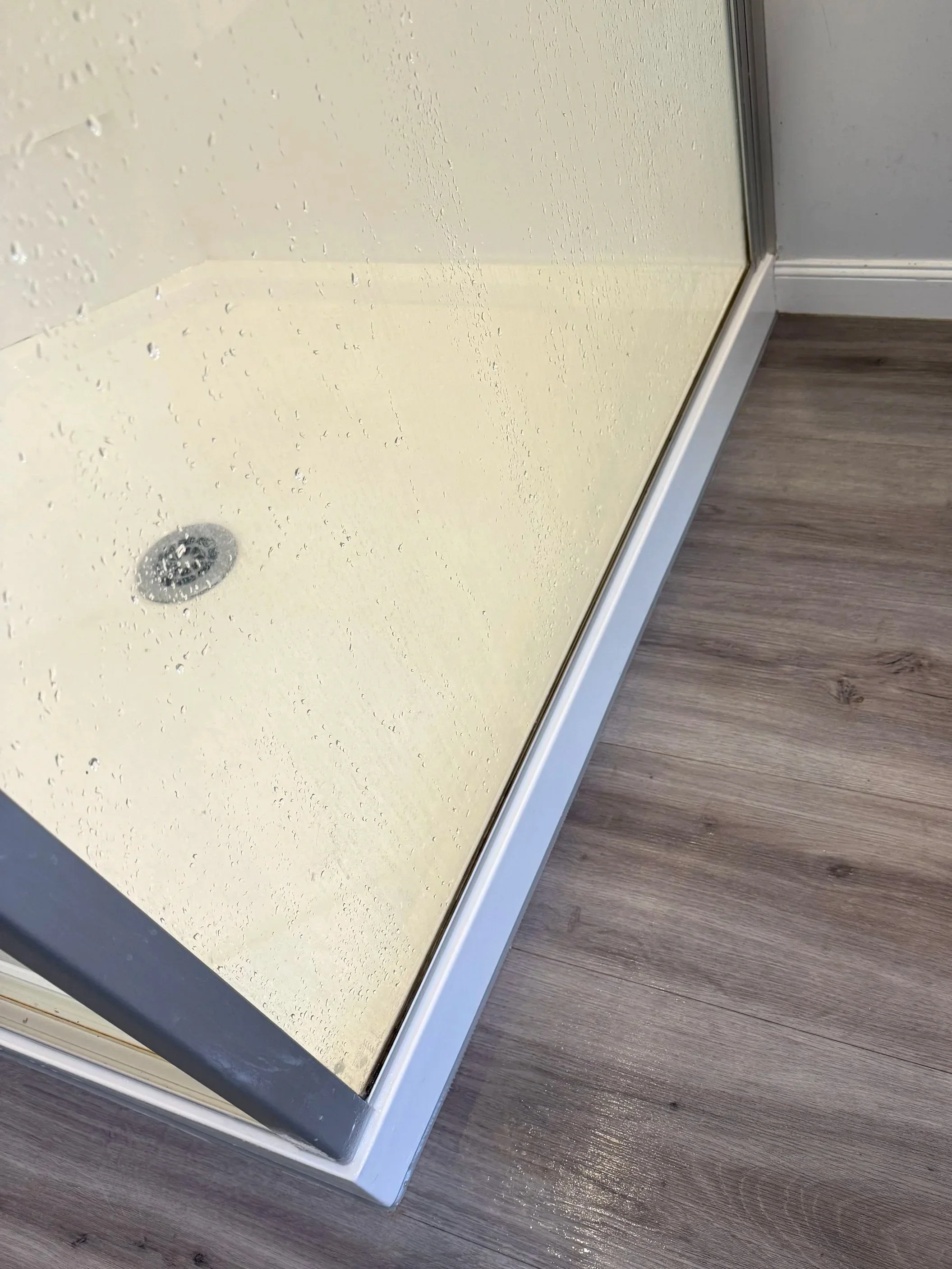 Water droplets on the glass shower door with a matte cream shower wall behind it, tiled wooden floor outside the shower.