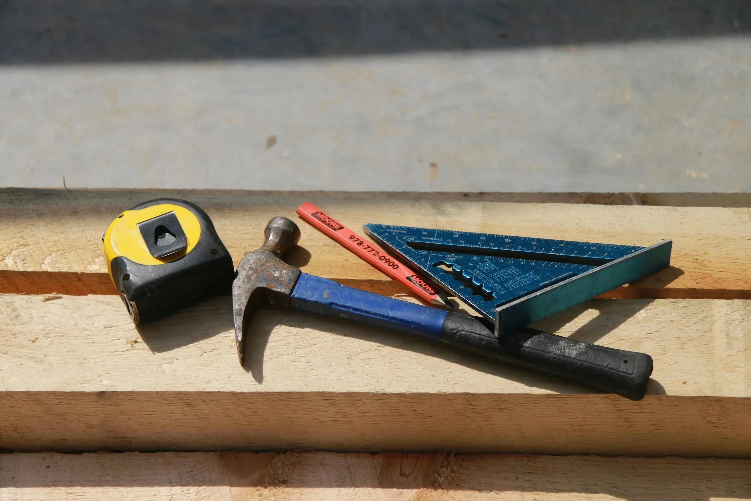 Construction tools including a measuring tape, hammer, speed square, and level resting on a piece of wood.