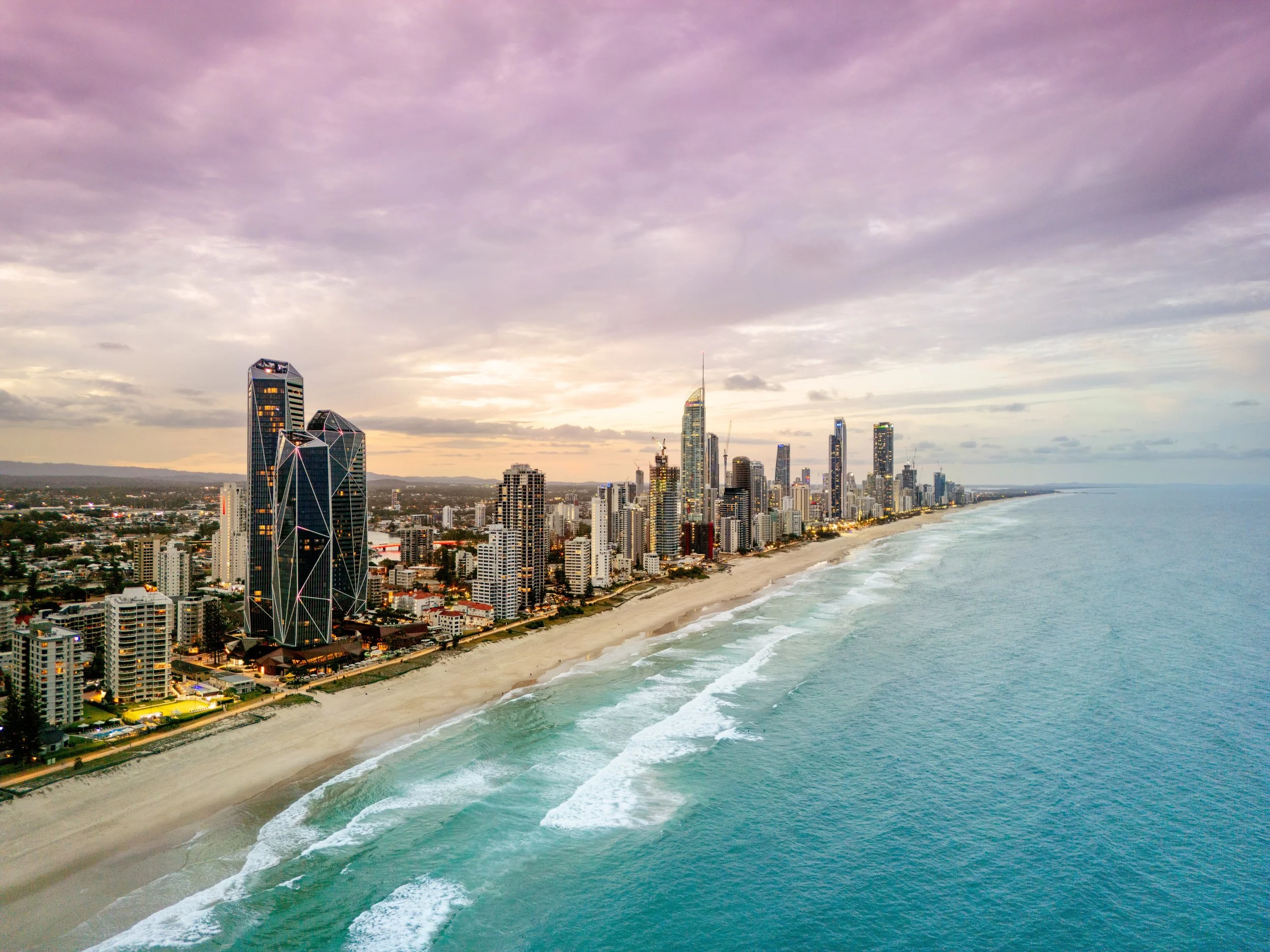 Modern office building with large glass windows and vertical beige panels, part of a city skyline with other tall buildings in the background.