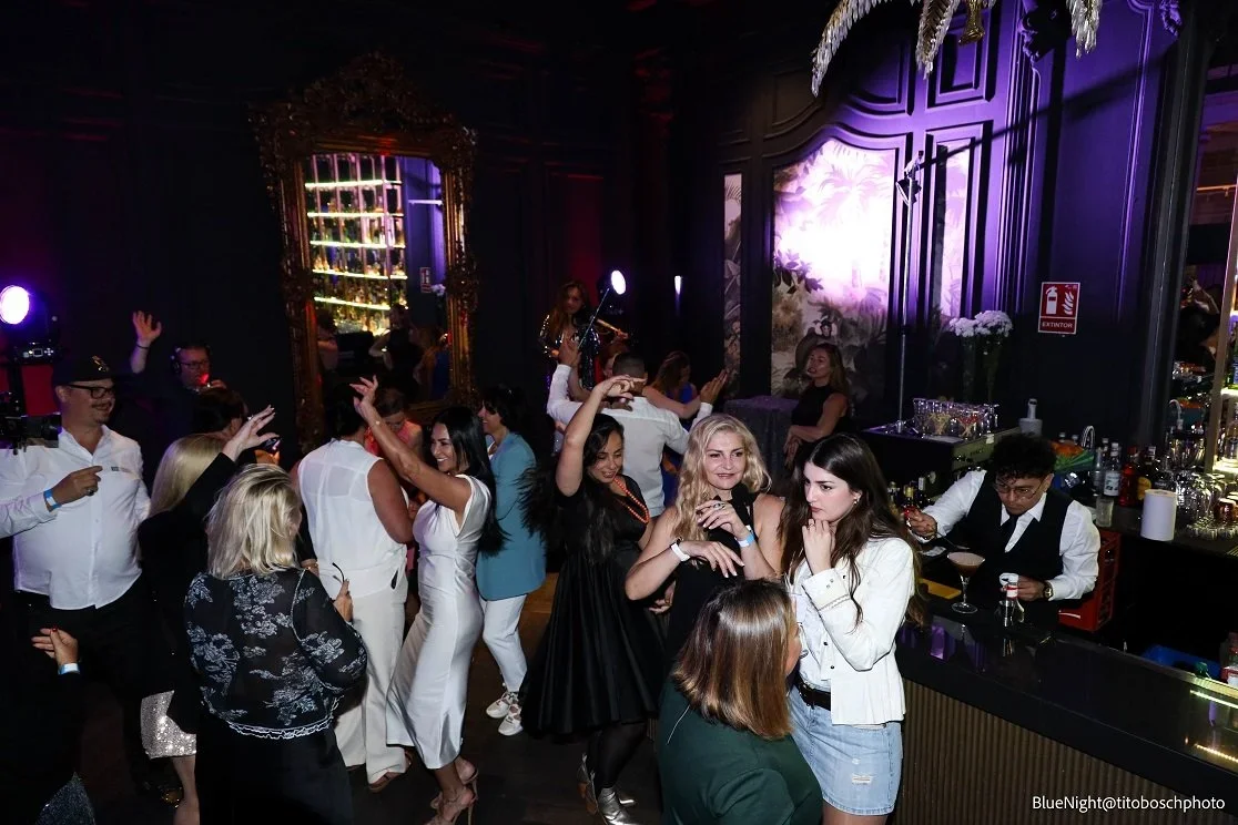 People dancing and socializing in a nightclub with colorful lighting and a bar area.